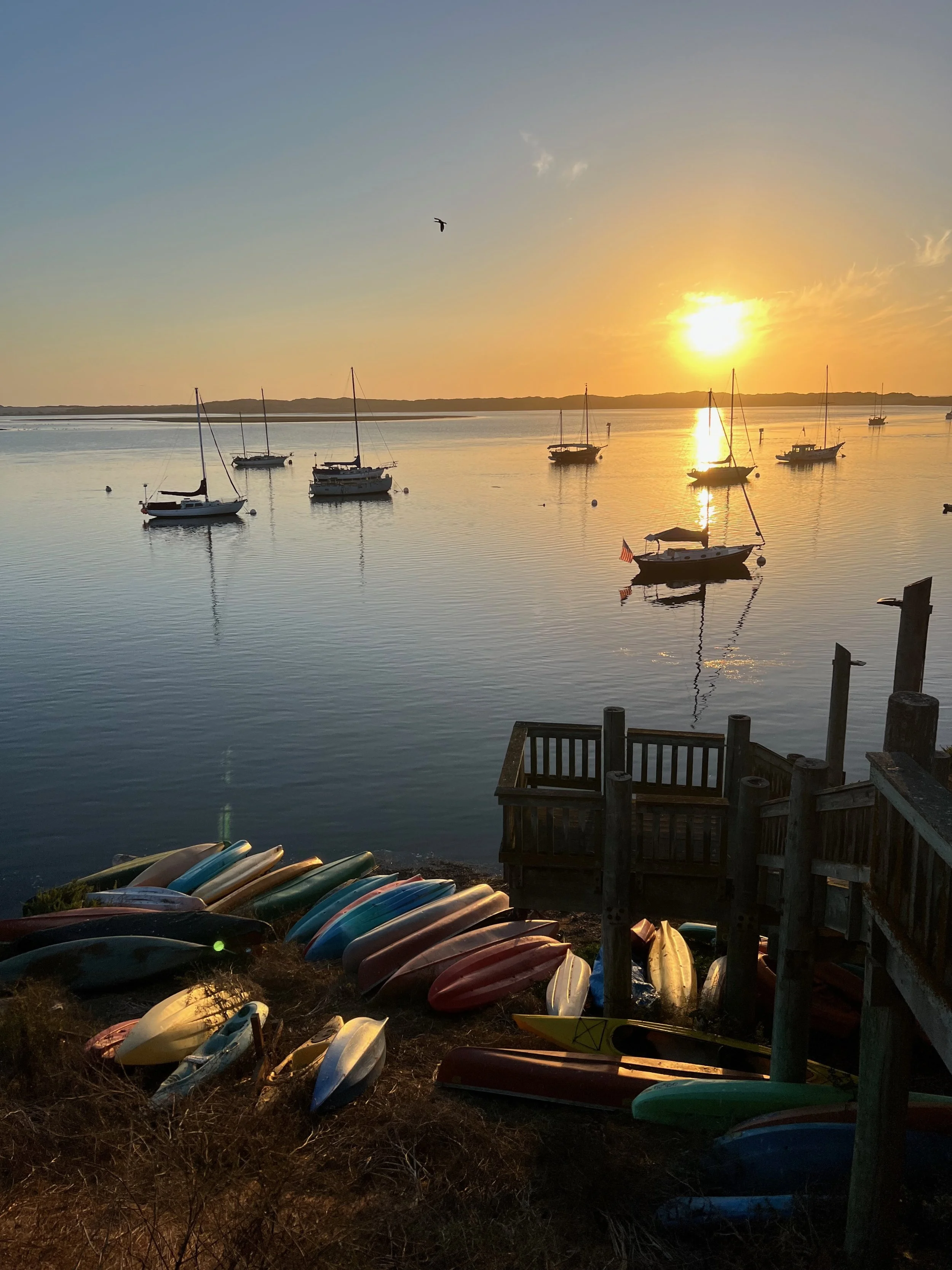 Sunset over a calm Morro Bay with sailboats anchored in the water and colorful kayaks piled on the shore | Tara Stephenson