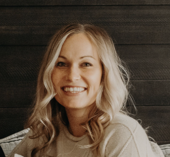 A smiling woman with blonde hair wearing a light-colored sweater, sitting in front of a dark wooden wall.