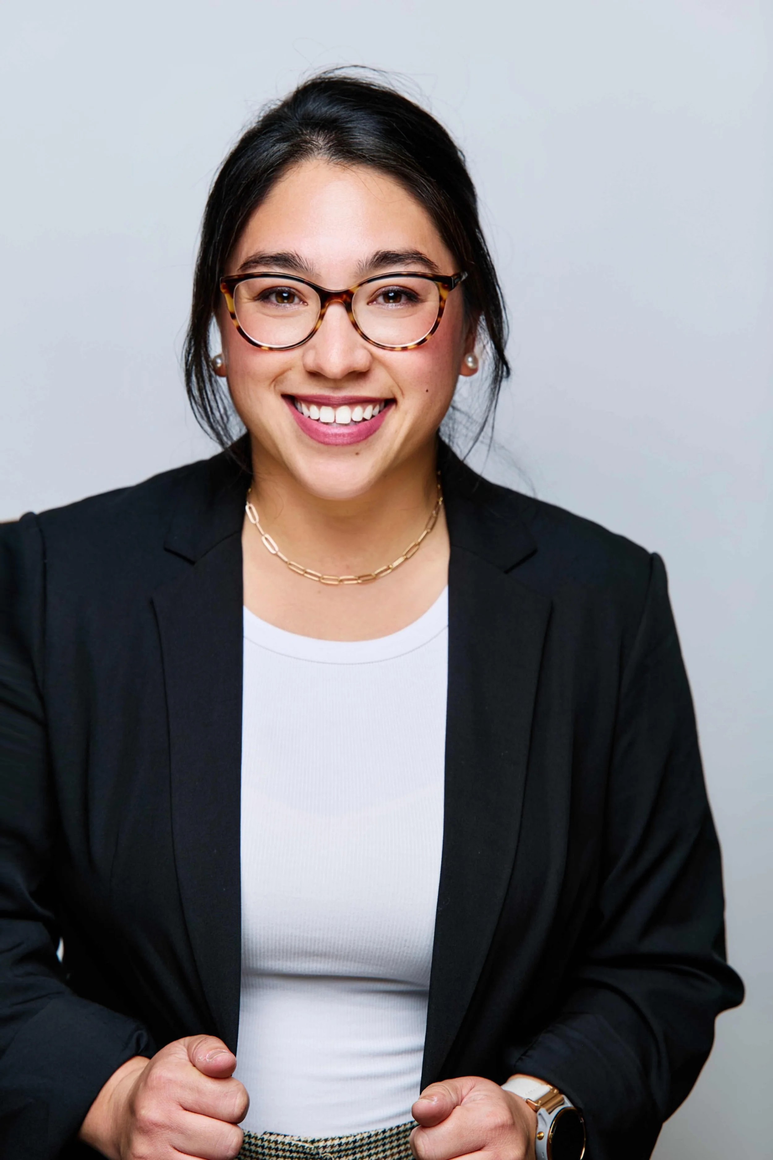 A young woman smiling, wearing glasses, a black blazer, a white shirt, and jewelry, standing against a light-colored background.