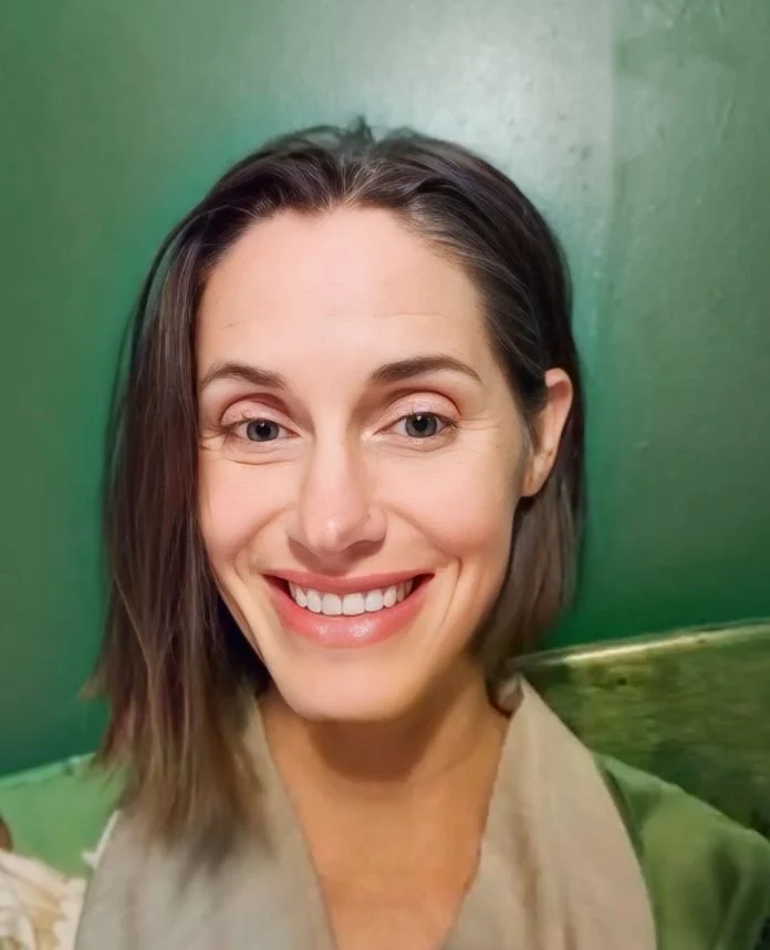 Smiling woman with shoulder-length brown hair standing in front of a green wall.