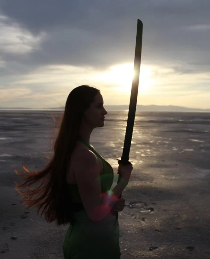 A woman with long hair holding a sword, standing on a beach during sunset, with the sun behind her and dark clouds overhead.