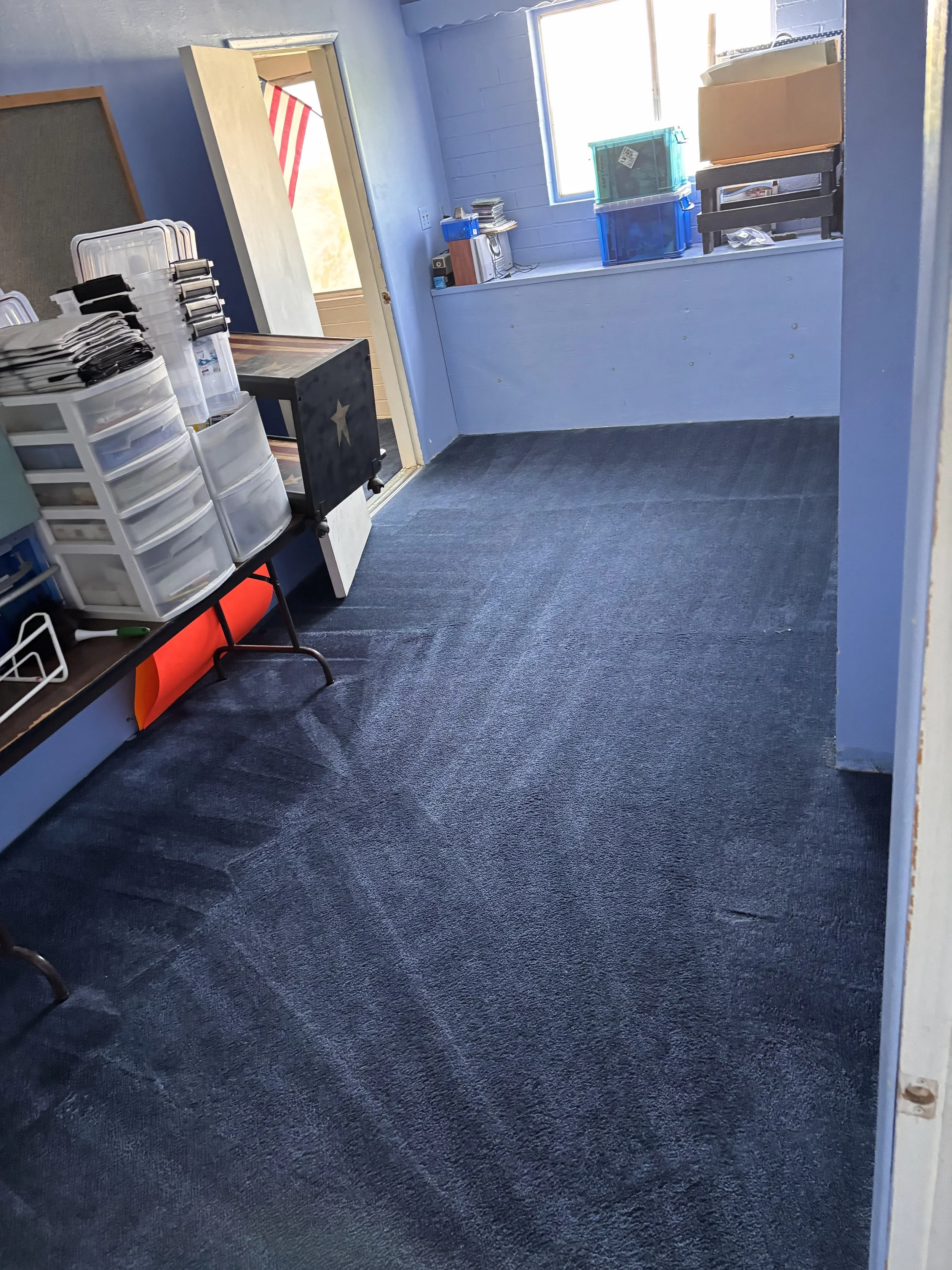 A room with light blue walls, a window with sunlight, a dark gray carpet, stacked plastic storage drawers, a small table with items on it, and some plastic containers and cardboard boxes near the window.