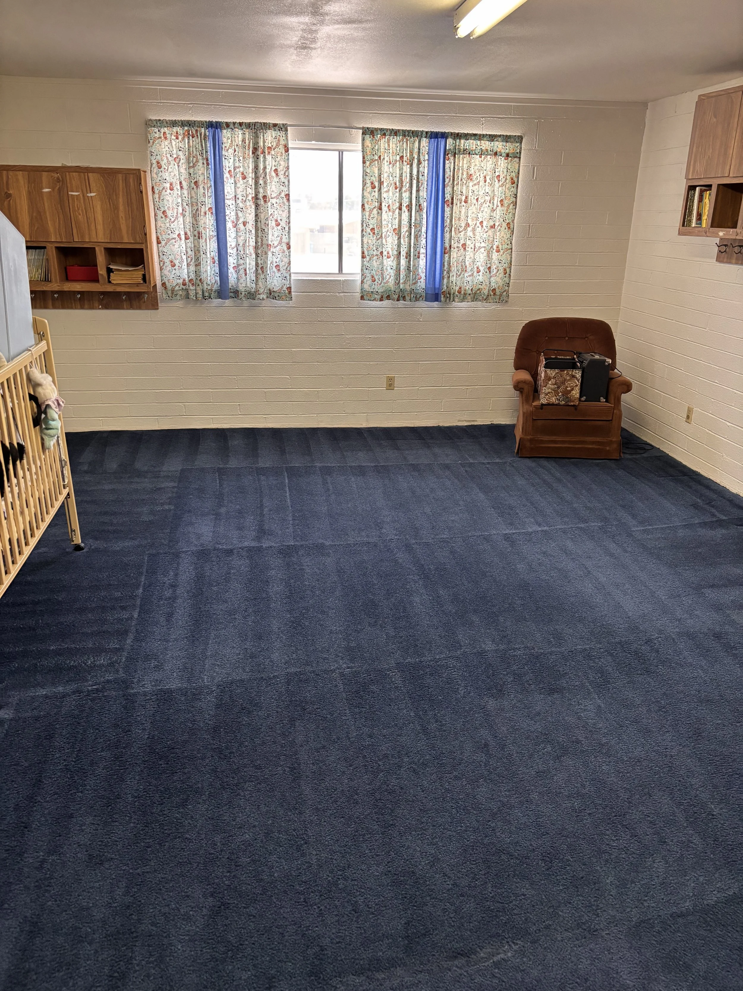 Photo of a room with white brick walls, blue carpet, a window with floral curtains, an armchair with items on it, and wooden shelves on the walls.