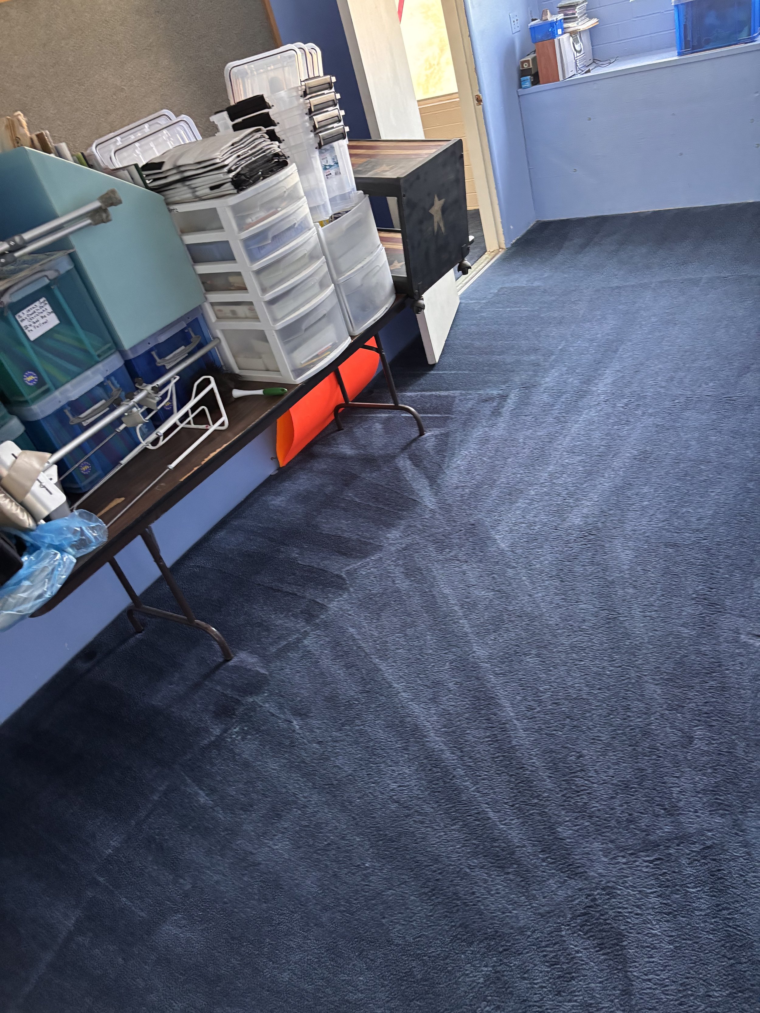 A classroom or office corner with a table holding organized plastic storage drawers and office supplies, a black cube with a star, and a wall with a window, blue wall, and carpeted floor.