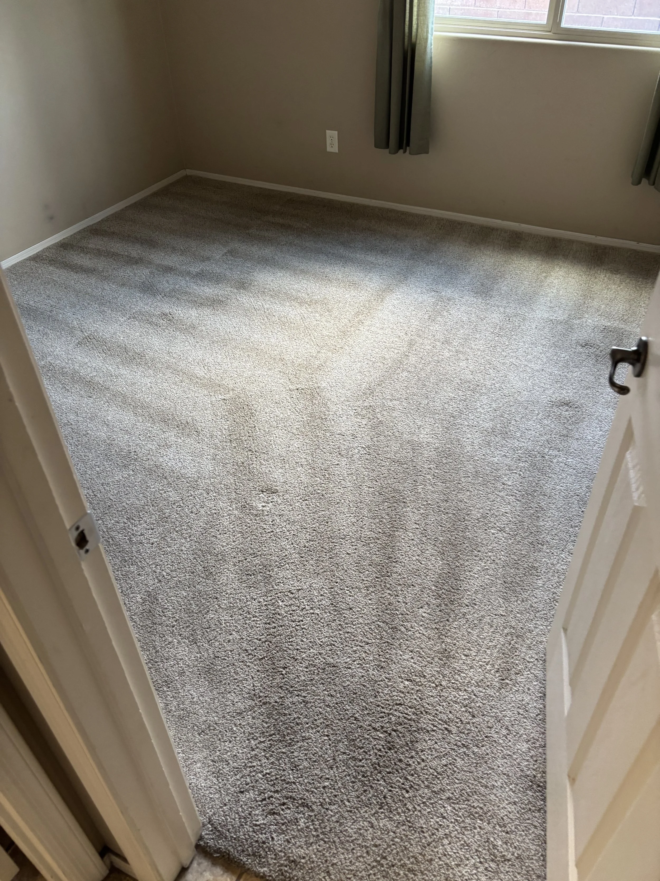Empty room with beige walls, brown and white speckled carpeting, a window with gray curtains, and a white electrical outlet on the wall.