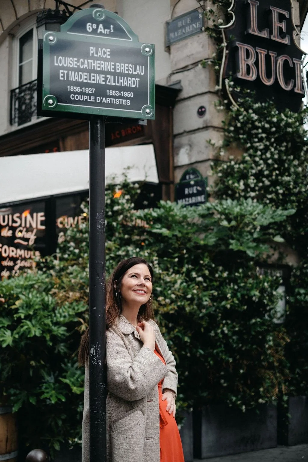 woman leaning against a street sign pole on a Paris street, with greenery and shop signs in the background.
