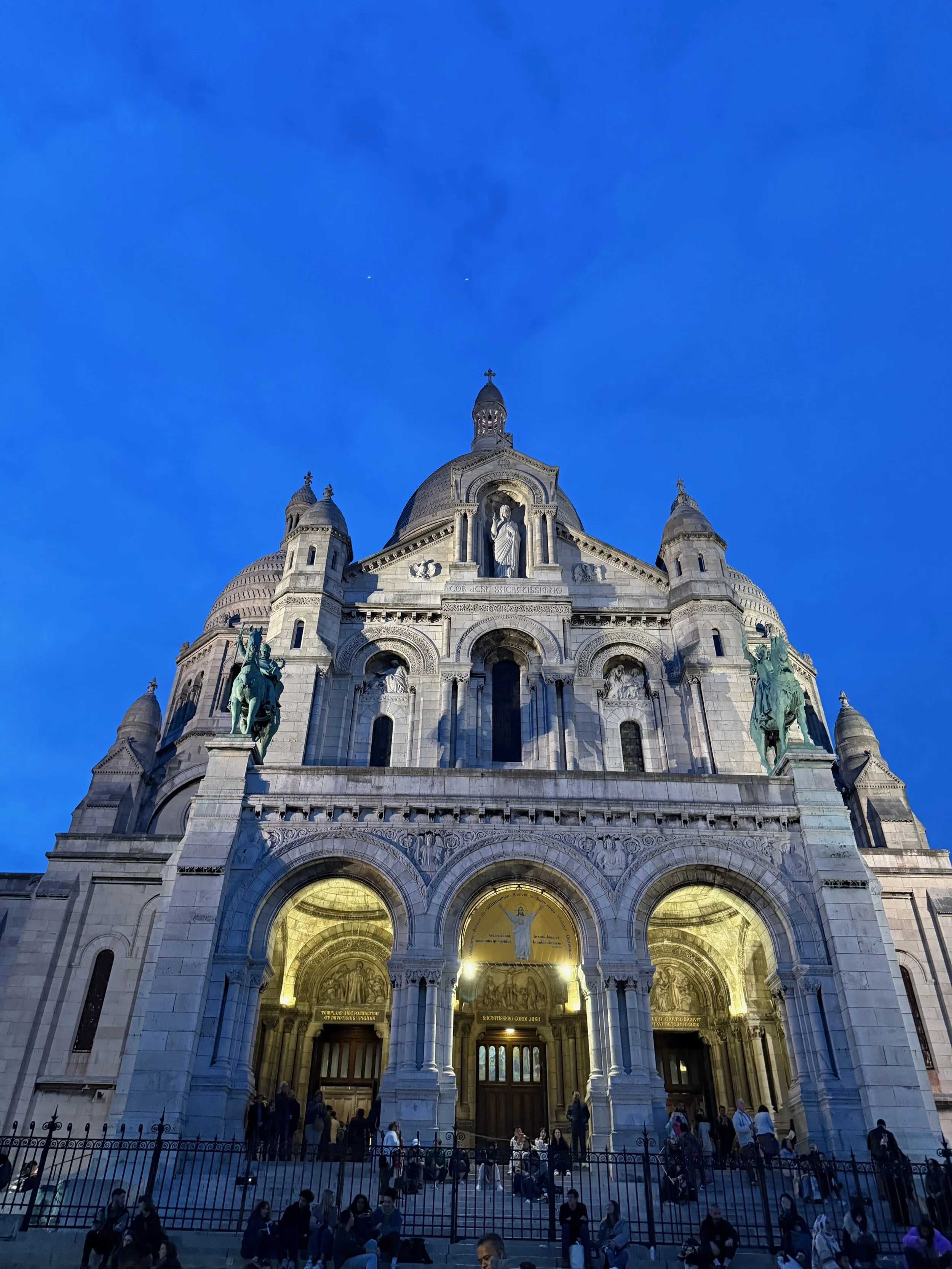 The Sacré-Cœur Basilica with stairs filled with people in front, illuminated at dusk, under a sky transitioning from blue to darker shades.