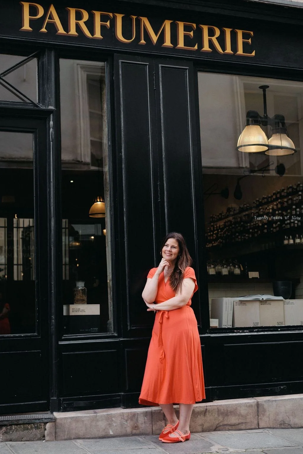A woman in an orange dress and matching shoes standing outside a black storefront that reads 'Parfumerie'. She is smiling with her hand resting on her chin.