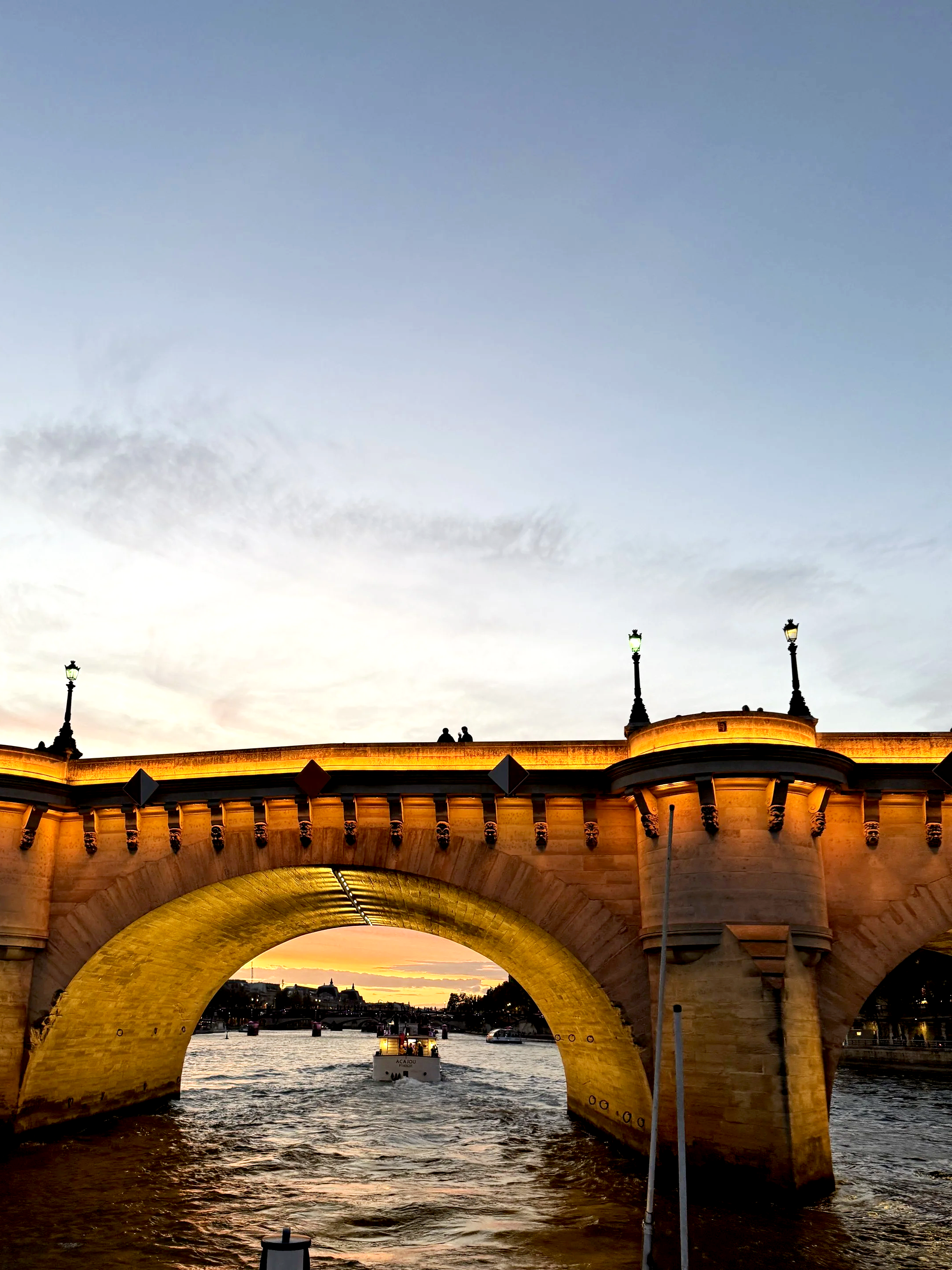 A view of a river under a bridge in Paris during sunset, with a boat passing through the arch of the bridge, and street lamps illuminated on top of the bridge.