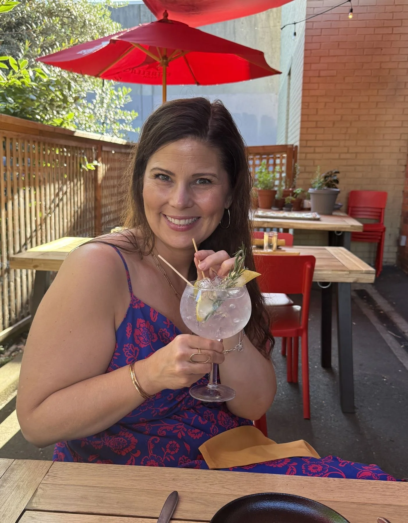 A woman with long dark hair, wearing a blue dress with red floral pattern, is smiling and holding a large glass of a clear cocktail with ice, lemon, and rosemary, at an outdoor restaurant with red chairs, wooden tables, potted plants, an umbrella, an
