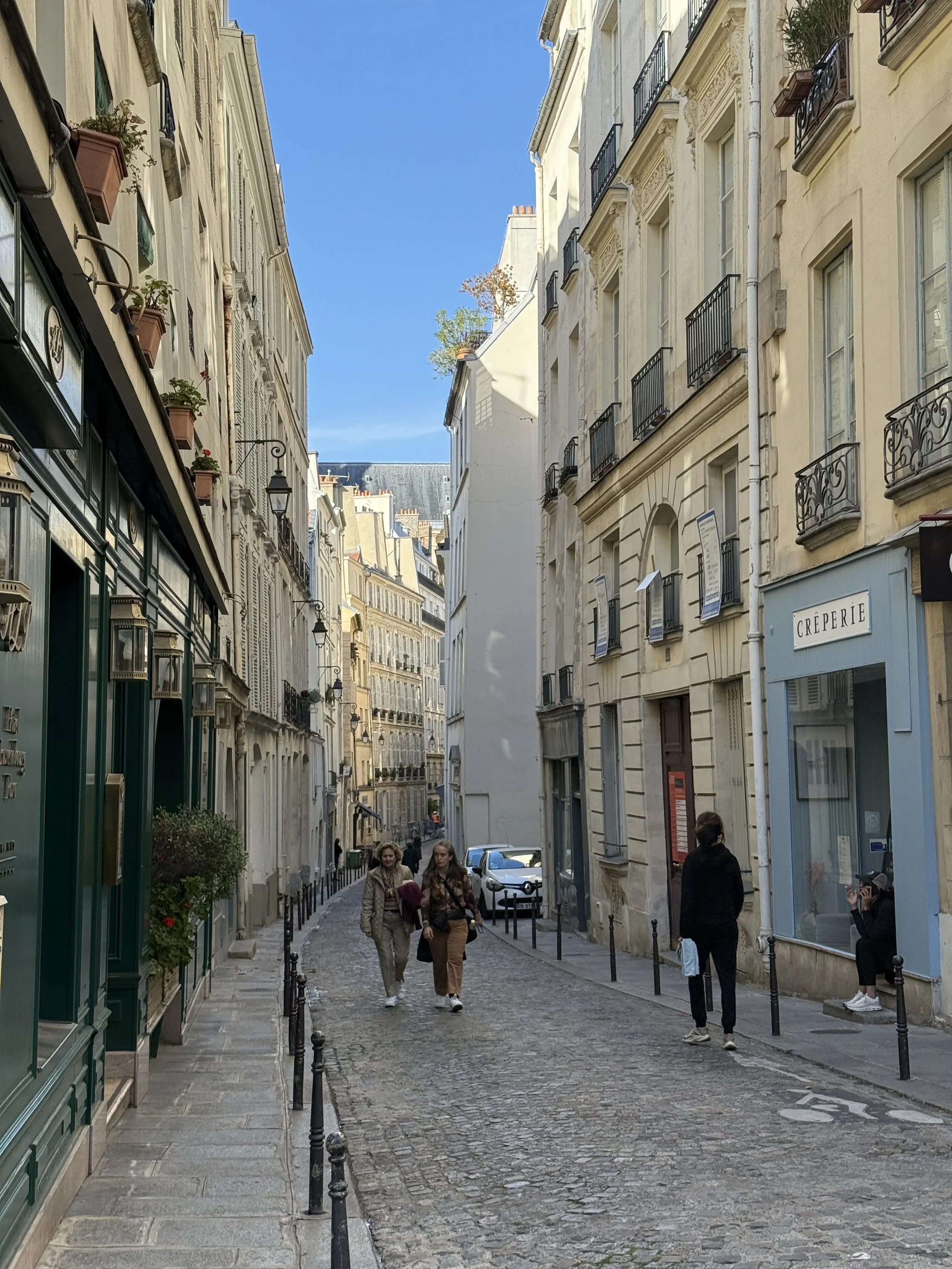 A narrow European street with cobblestone pavement, lined with cream-colored multi-story buildings with balconies, windows, and street lamps. Three women walk down the street, and one person is taking a photo.