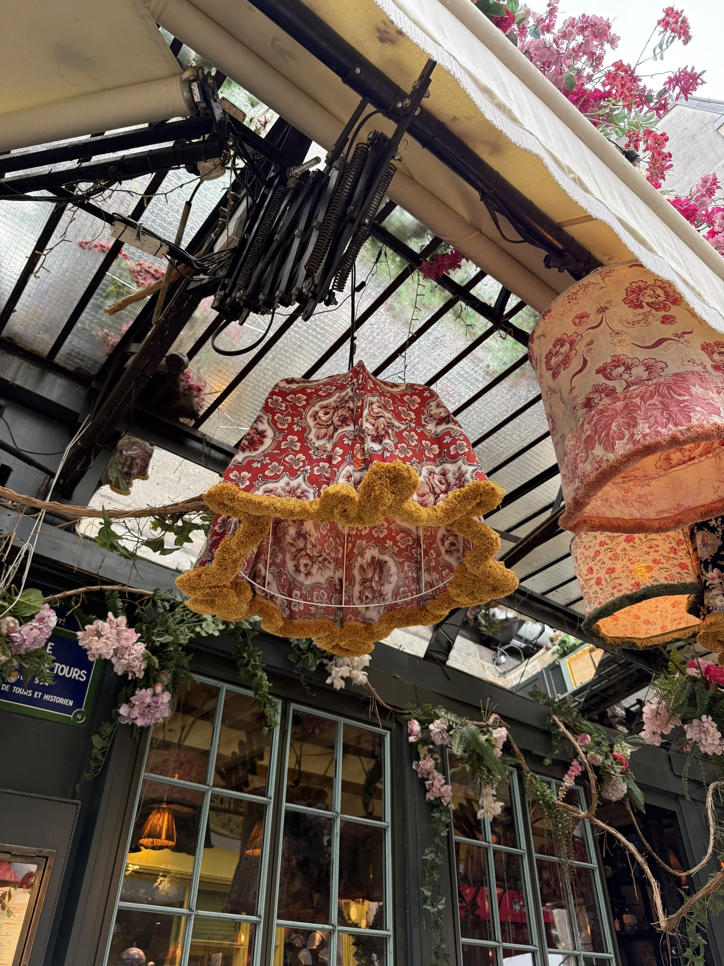 Colorful hanging lampshades, including one with floral fabric, are suspended outside a building with glass windows. Pink flowers and green vines adorn the area, creating a vibrant and decorative atmosphere.