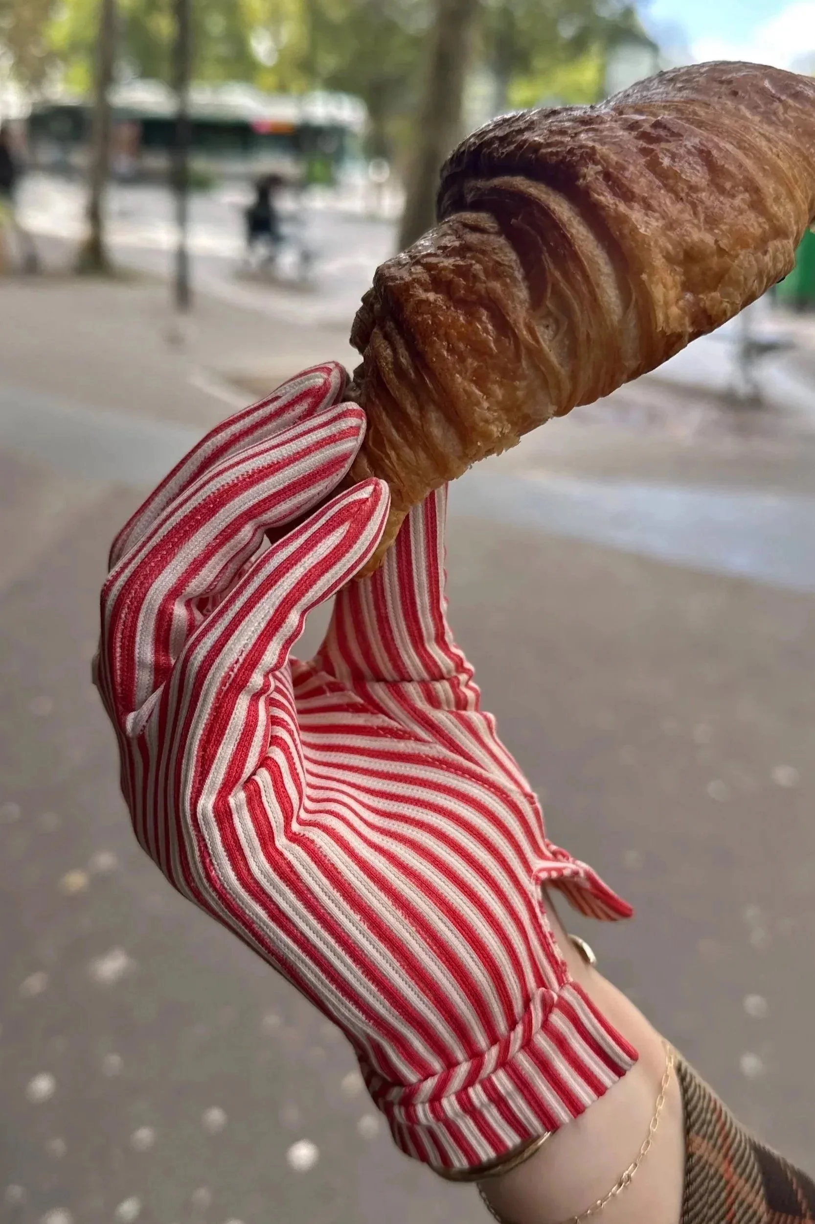 A person's hand, wearing red and white striped gloves, holds a croissant with a background of trees and blurry pedestrians in an outdoor setting.