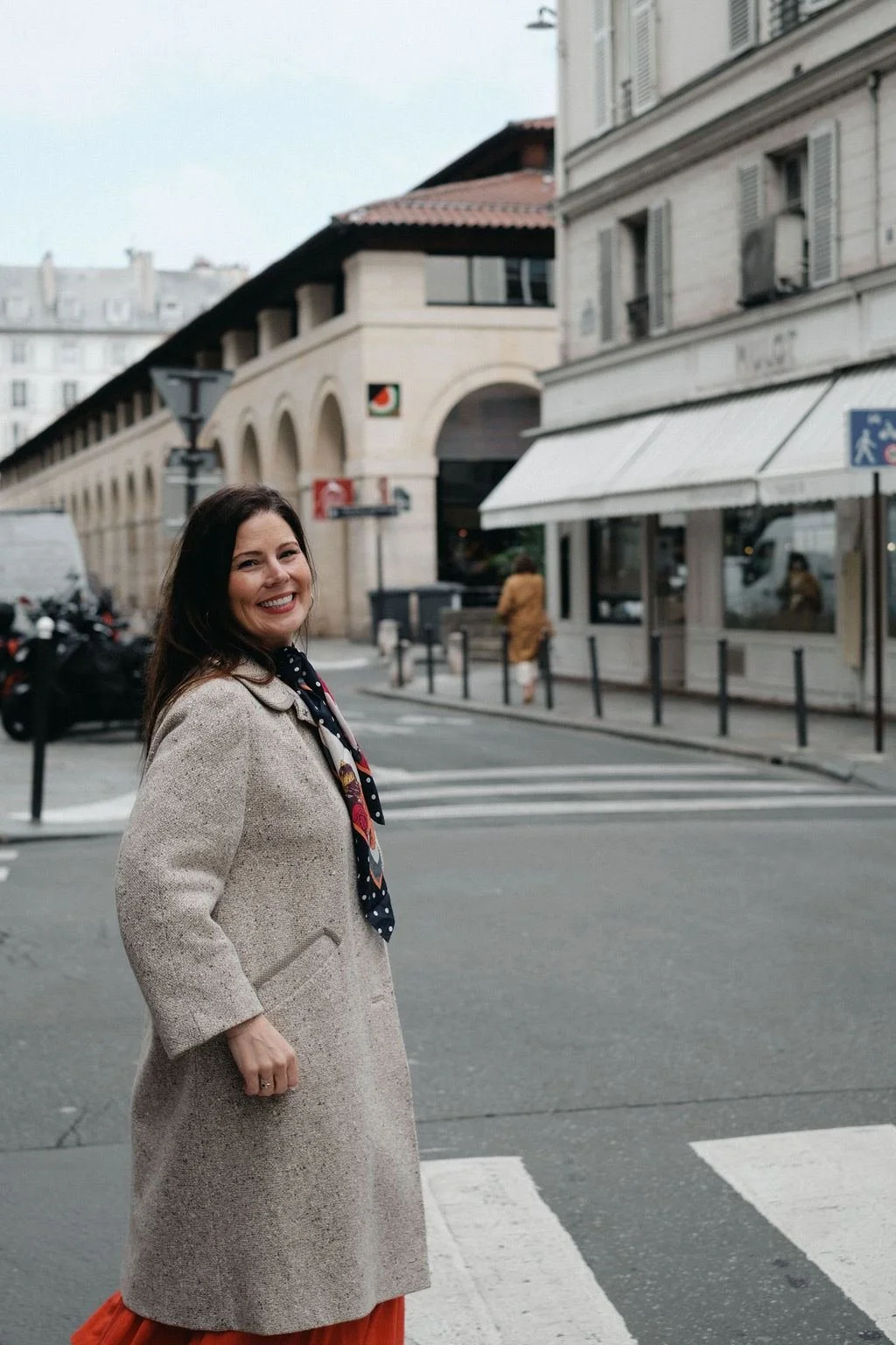 A woman with dark hair, smiling, standing on a crosswalk in an urban street with buildings and pedestrians in the background.