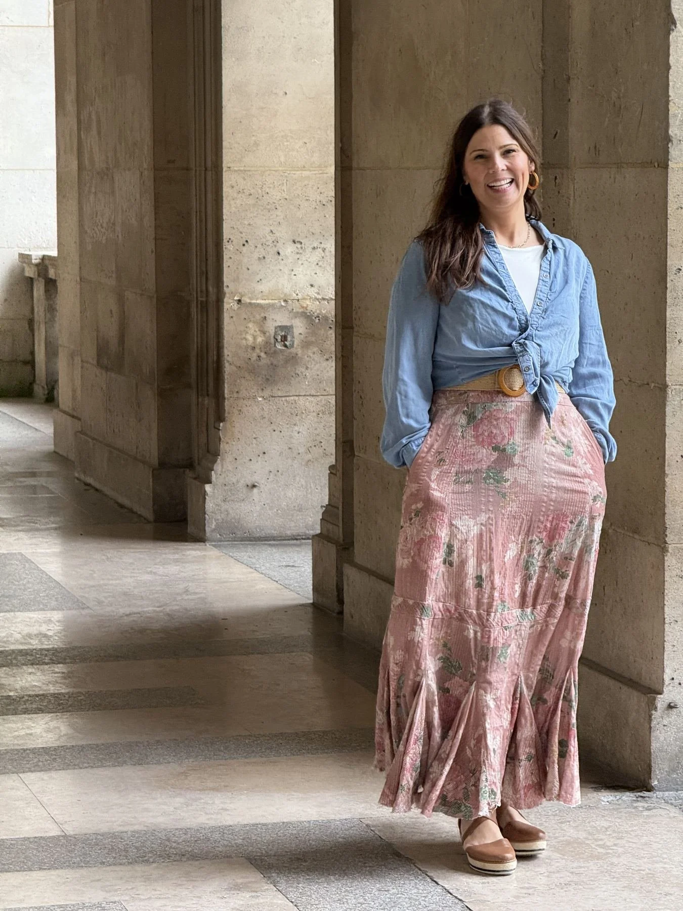 A woman stands in a hallway with beige stone walls and a tiled floor, smiling and wearing a light blue denim shirt tied at the waist, a floral pink long skirt, and beige platform shoes.