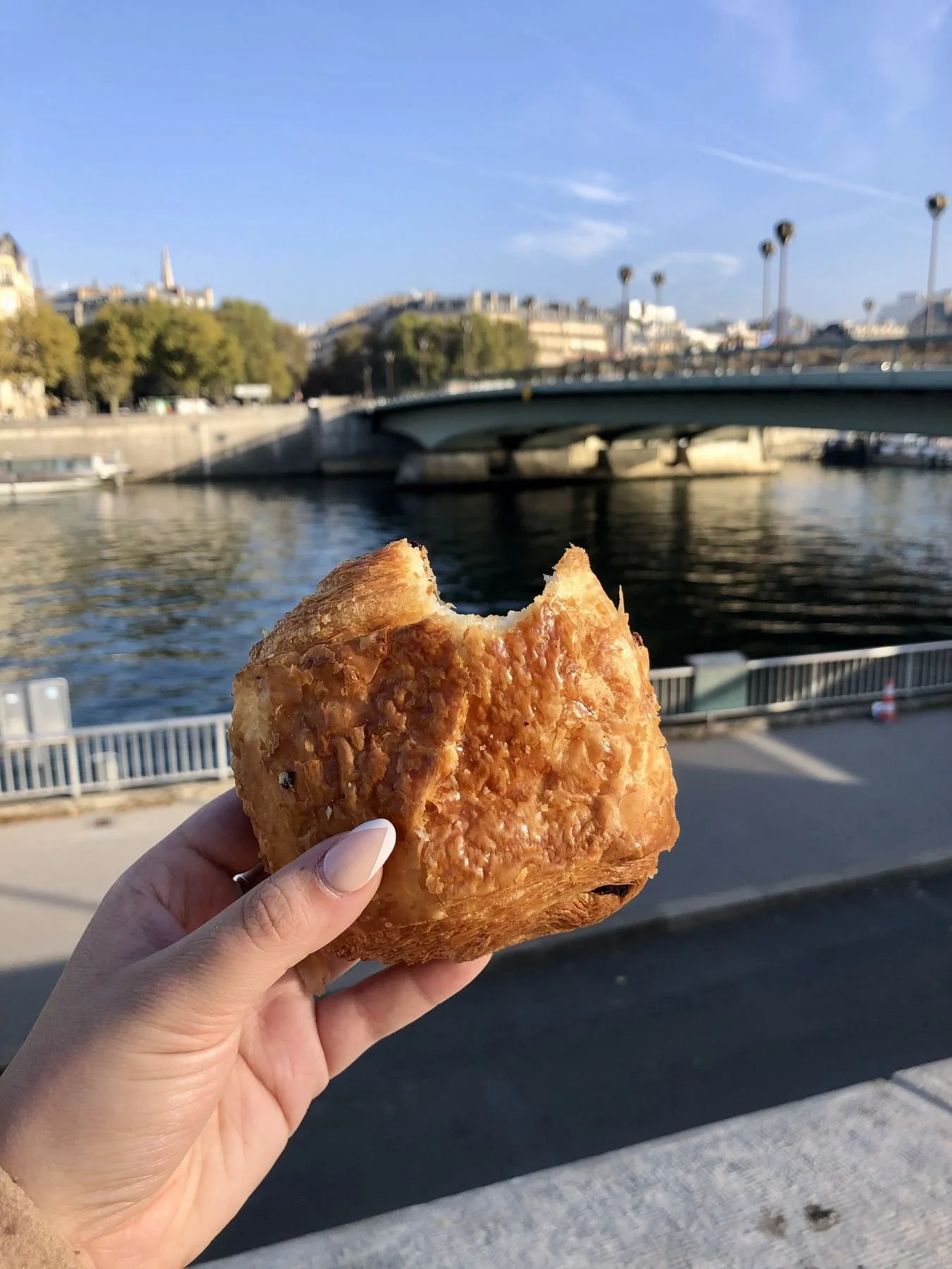 A person holding a piece of fried pastry or turnover near a river, with a city bridge and buildings in the background on a clear day.
