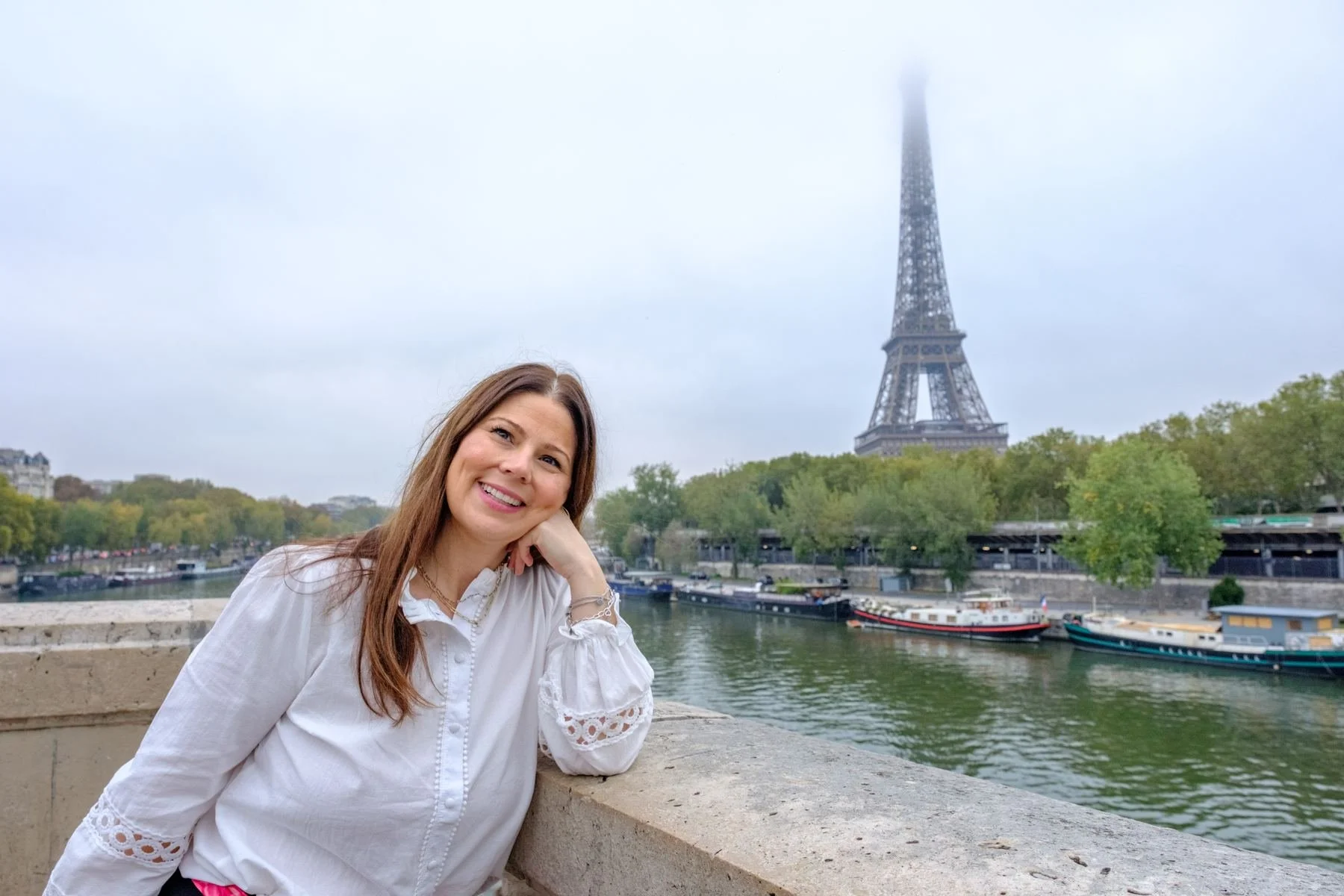 A woman with long brown hair, wearing a white blouse with lace details, is sitting by the Seine River in Paris, with the Eiffel Tower shrouded in fog in the background.