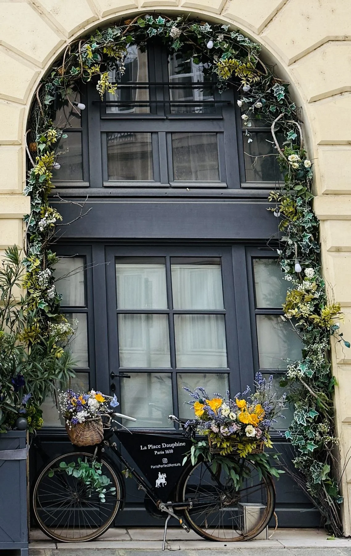 A black bicycle with a wicker basket on the front, decorated with a floral arrangement, parked in front of a building with dark blue French doors and windows, surrounded by green ivy and yellow flowers.