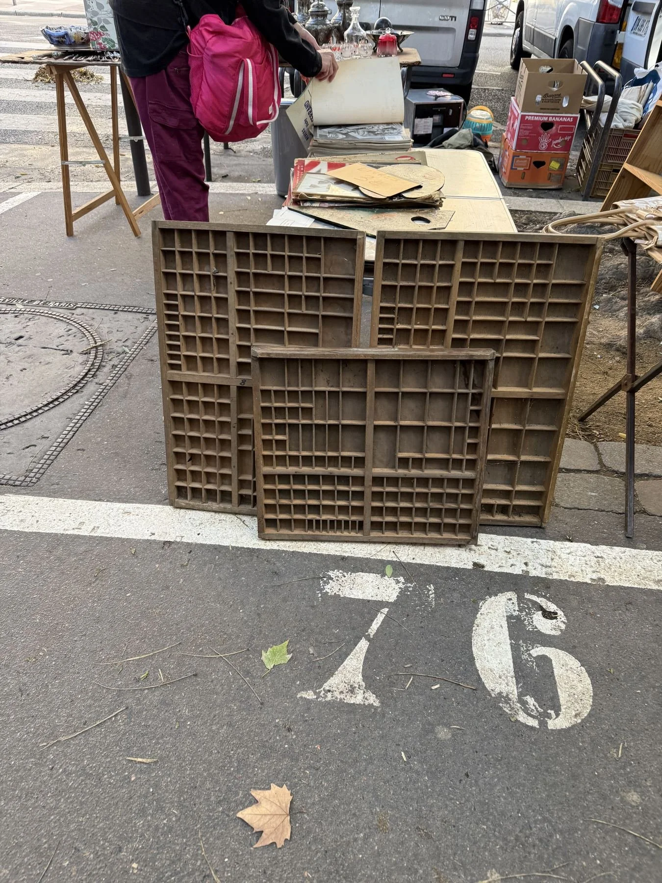 A flea market stall set up on the sidewalk with various items, including cardboard boxes, papers, and glassware, with a person browsing at the stall. The foreground shows white parking space markings on the asphalt, with a few fallen leaves.