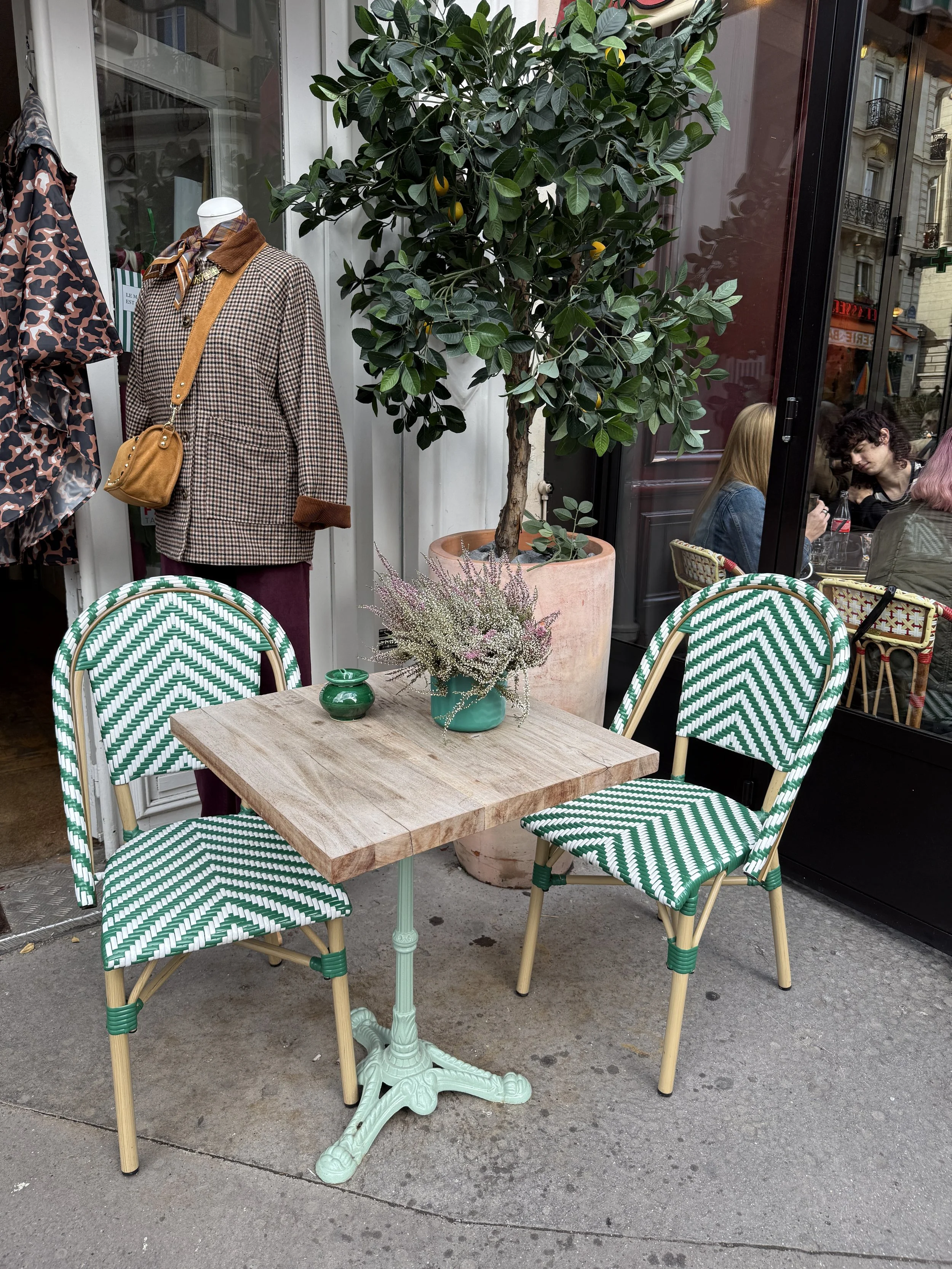 A small outdoor table with a green vase and a pinkish flower arrangement, surrounded by four green and white patterned chairs, outside a café with large windows and people sitting inside.