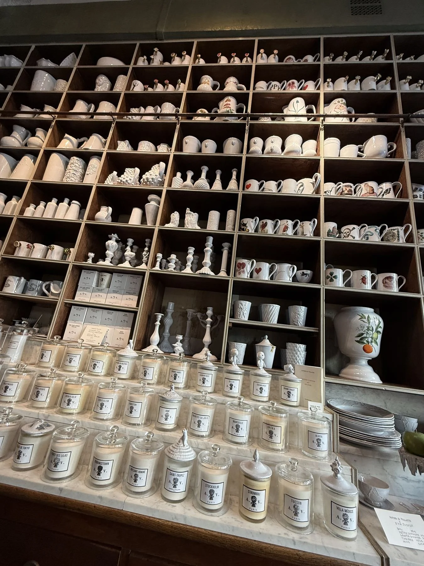 Display of white candles with labels of various city names in glass jars on a marble countertop, with shelves of white ceramic mugs and decorative items above.