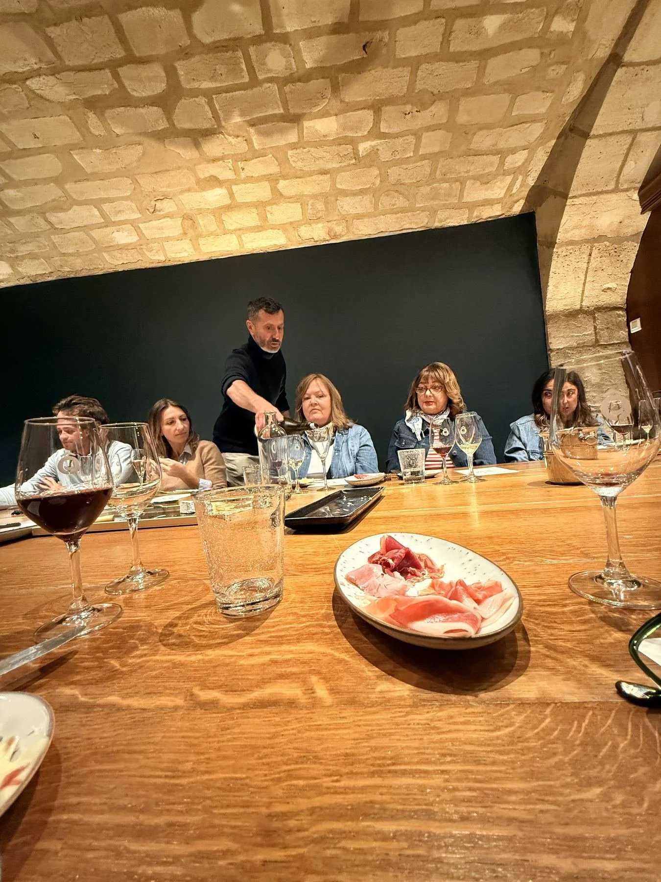 A group of people sitting at a long wooden table in a restaurant, with a man serving wine to a woman. The table has glasses of wine, water, and a plate of sliced meats. The background features a brick ceiling and a dark wall.