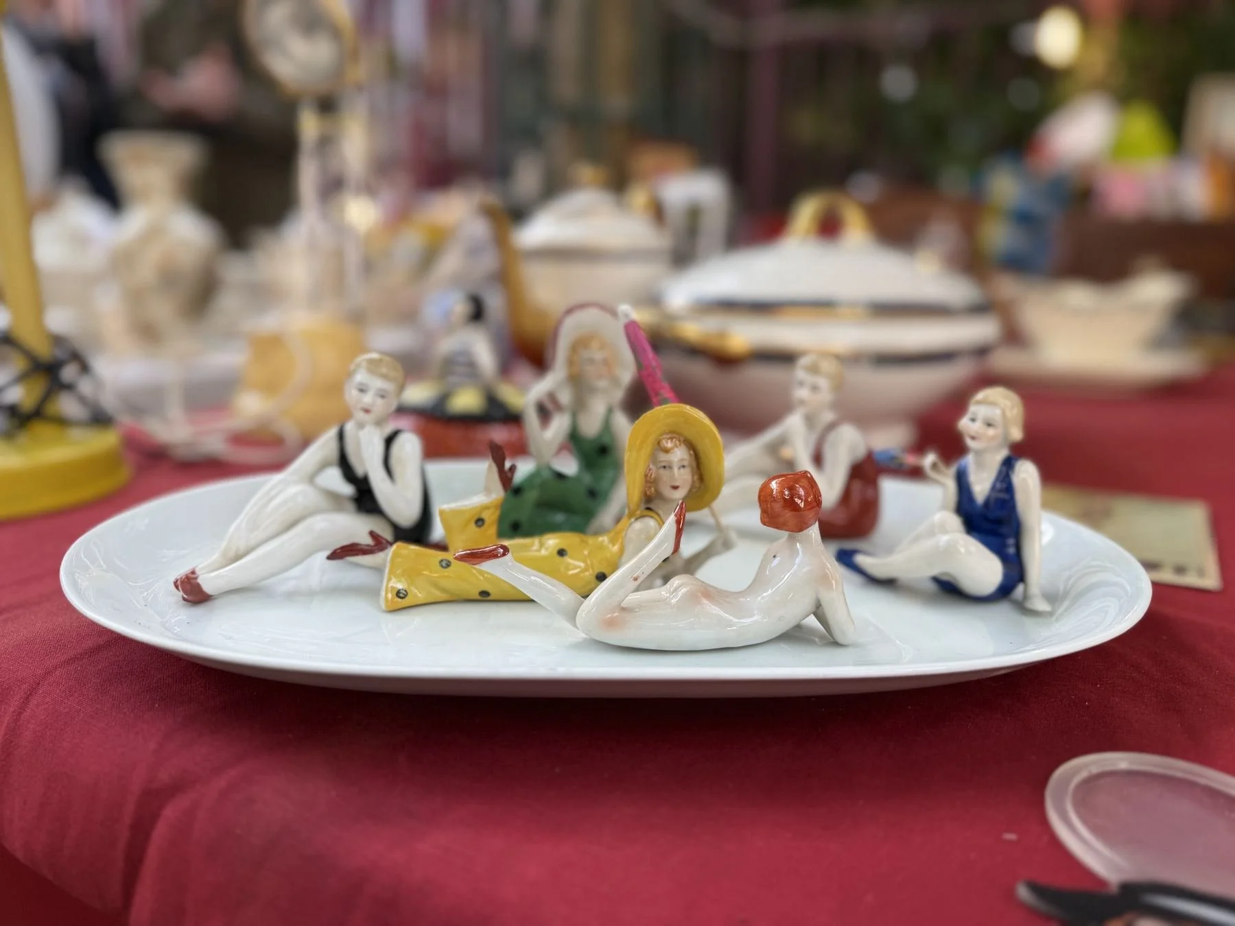 Decorative ceramic figurines of people and animals on a white plate, with more ceramic items and display tables in the background at a market or craft fair.