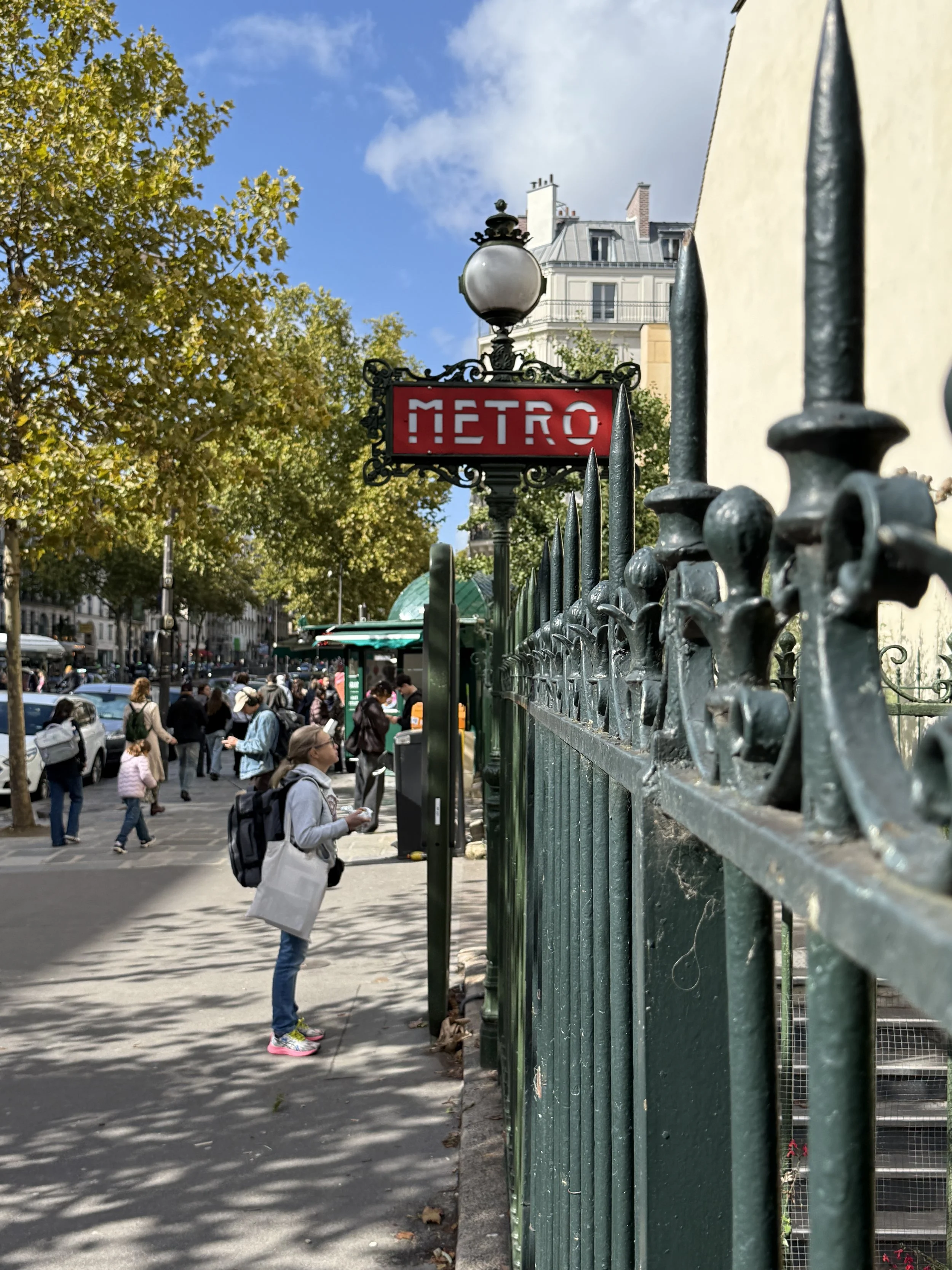 A crowded city sidewalk scene with people walking, a green metal fence in the foreground, a red 'Metro' sign, a lamppost, trees with green leaves, and Parisian-style buildings in the background under a partly cloudy sky.