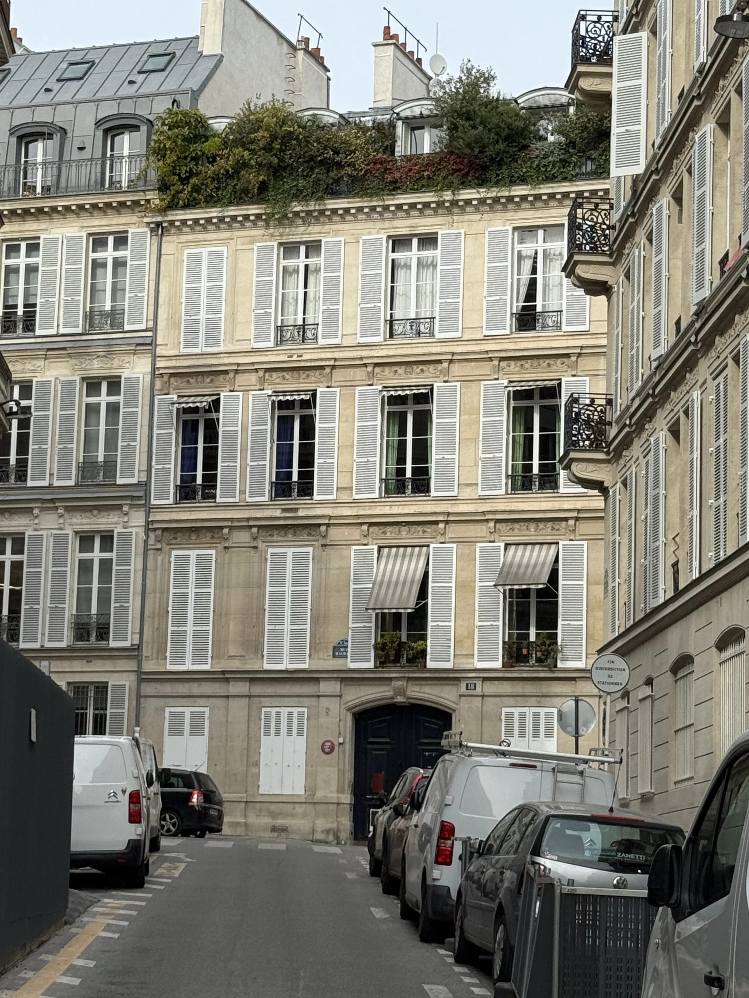 Street view of a residential building with multiple floors, white shutters on windows, and some greenery on the rooftop. Cars are parked along the street in front of the building.