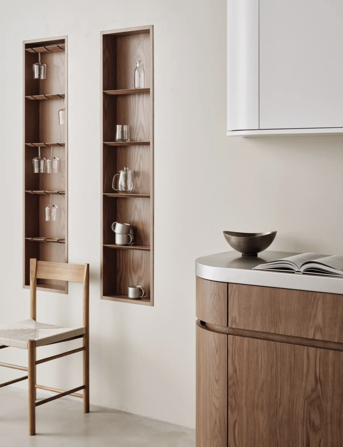 Minimalist kitchen with two wooden wall-mounted shelves displaying glassware and mugs, a wooden chair, and a curved wooden cabinet with an open book and a black bowl on top.