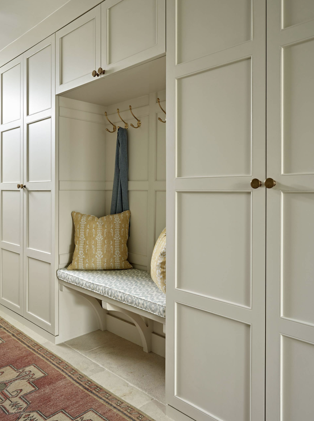 Built-in white closet with gold hooks, a cushioned bench with pillows, and a rug on the floor.