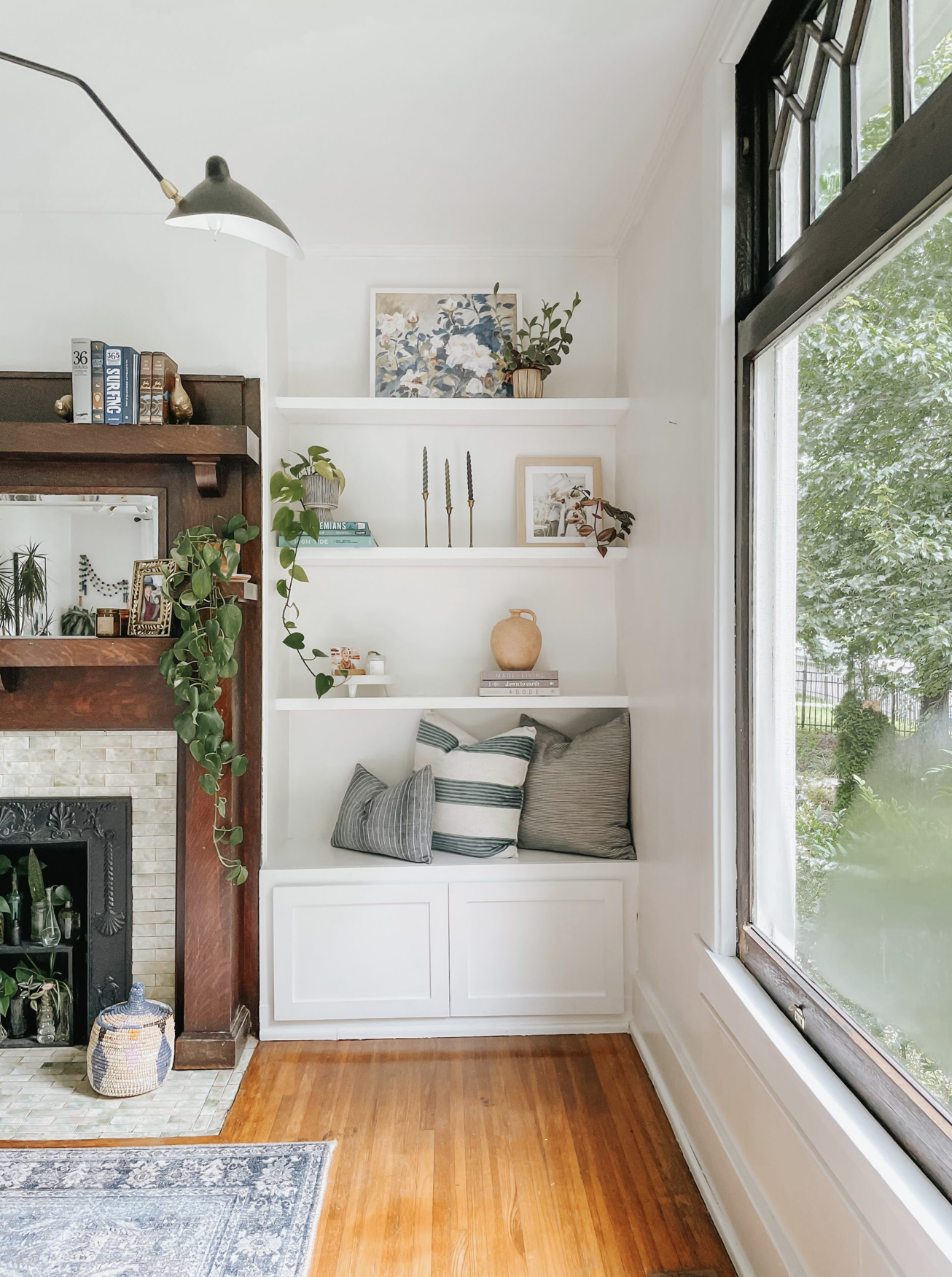 A cozy living room corner featuring white built-in shelves with decorative items and potted plants, adjacent to a large window with greenery outside, and part of a fireplace with a wooden mantel and decorative tiles.