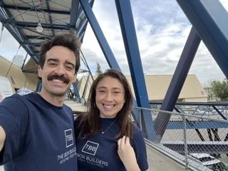 A man and woman taking a selfie outdoors under a metallic structure, both wearing matching blue T-shirts and smiling.