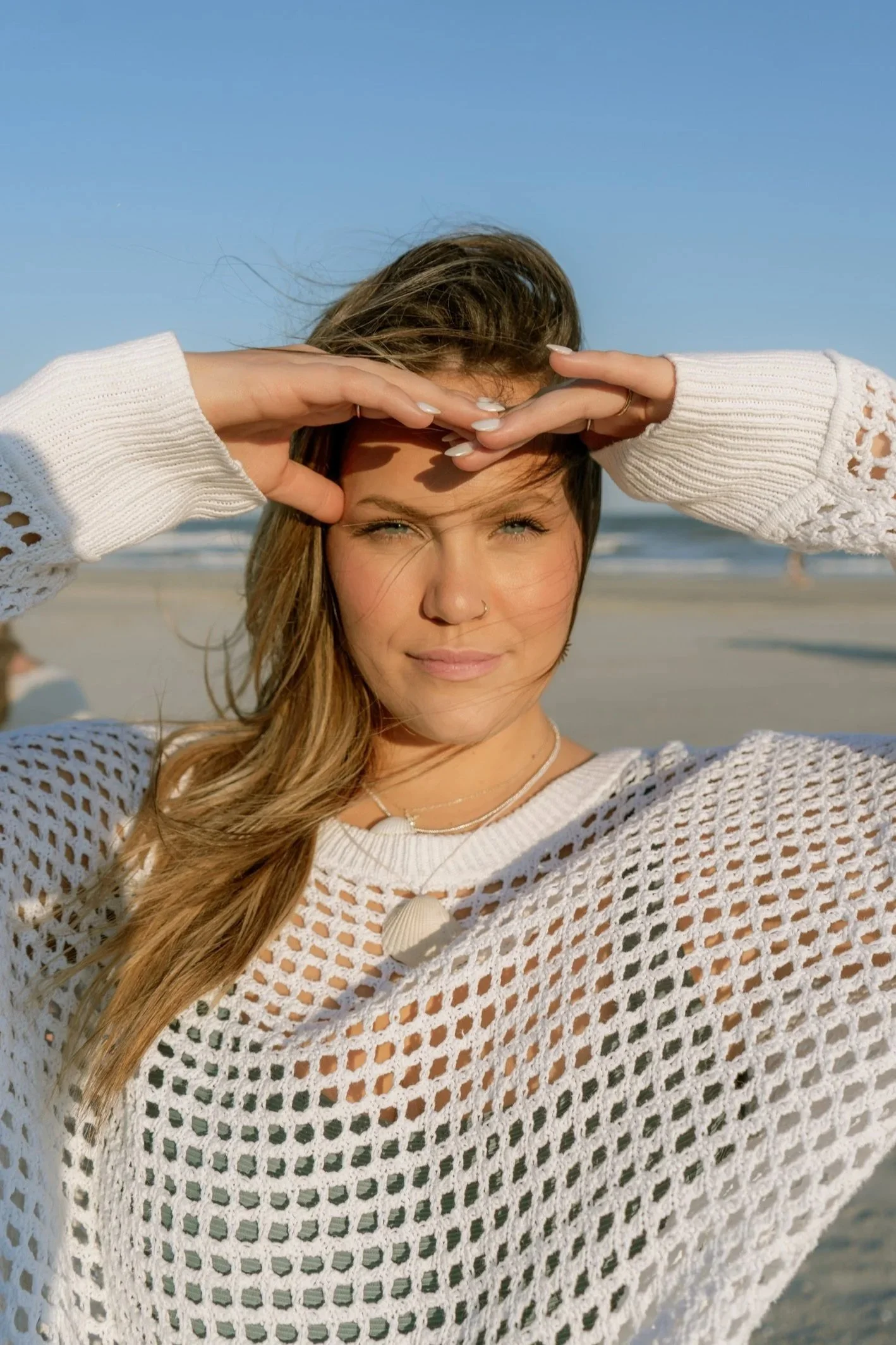 Woman at the beach shielding her eyes from the sun with her hands, wearing a white mesh sweater, with ocean in the background.