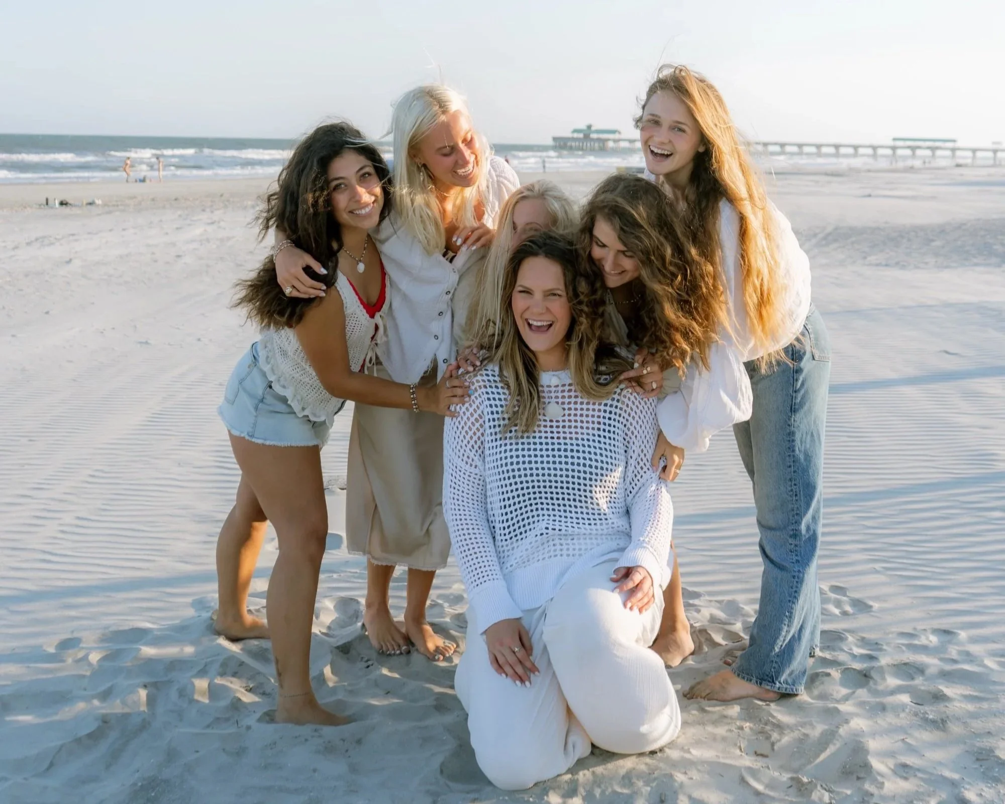 Group of six women laughing and enjoying at the beach, some kneeling and some standing on the sand with the ocean and pier in the background.