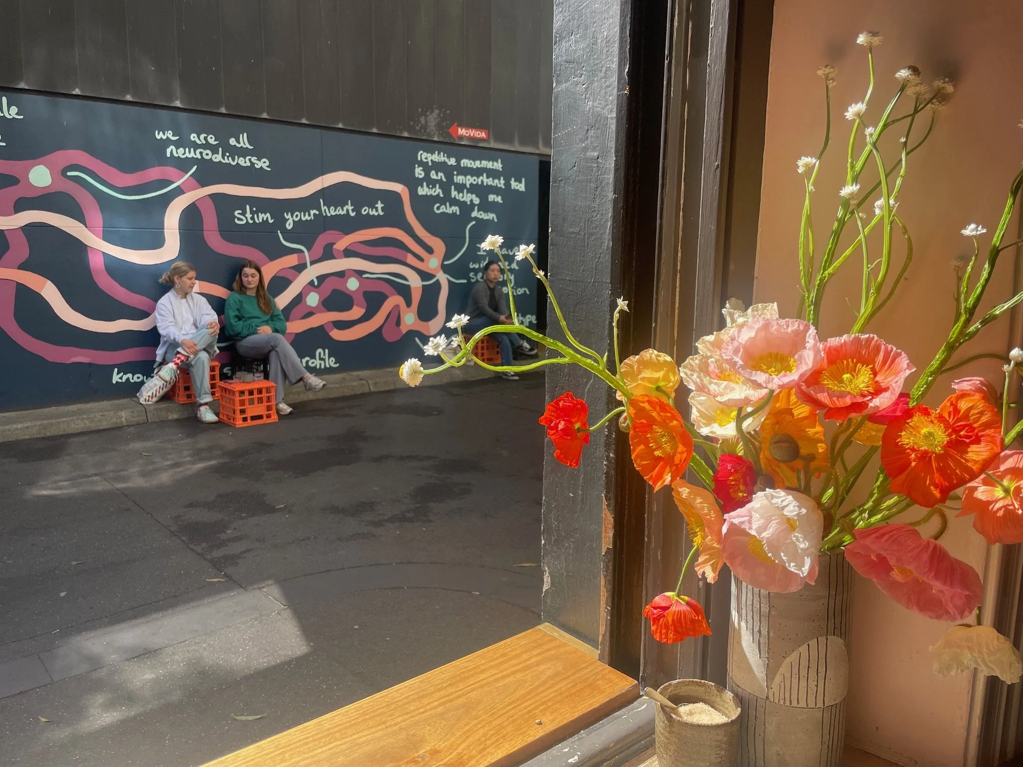 Looking out a window of the Melbourne cafe Patricia to the laneway outside. A vase of poppies is on the windowsill. Outside two woman sit on milk creates in front of a mural that says  "stim your heart out" and "we are all neurodiverse"