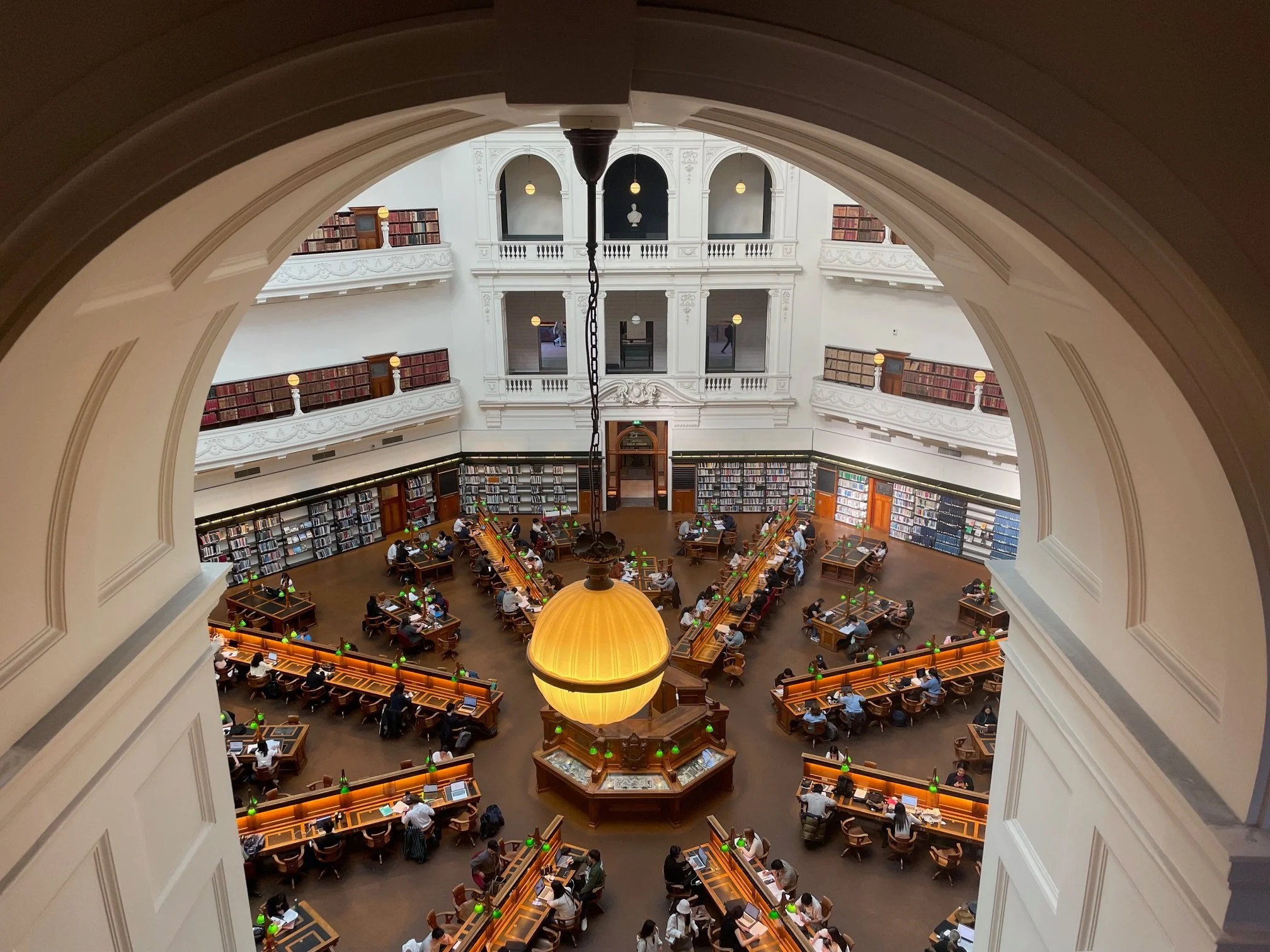 A view from the balcony looking down into the beautiful Dome Reading Room at the State Library of Victoria. People work at long desks in a hexagon shaped room lined with shelves of books.
