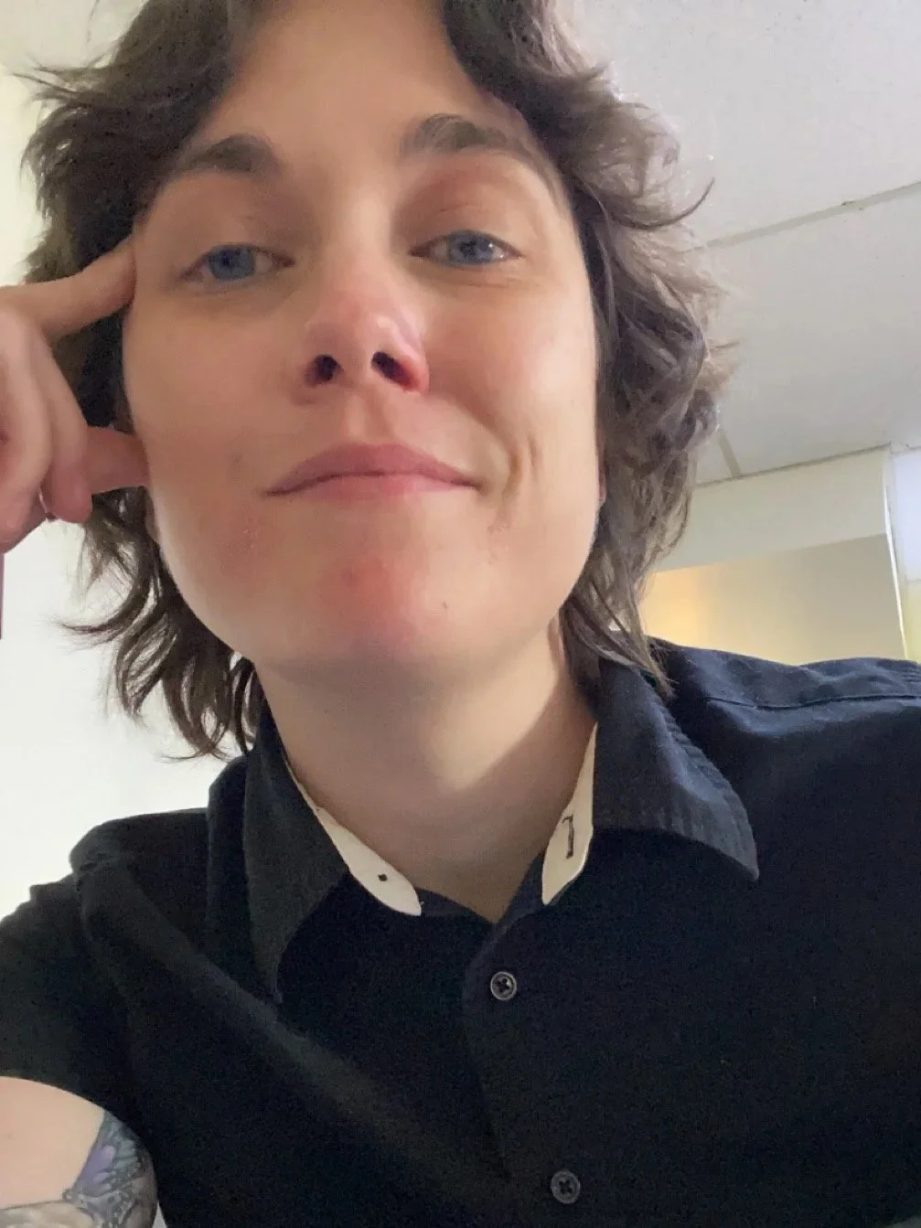 Close-up of a non-binary person with wavy brown hair, blue eyes, and wearing a black collared shirt with white details, resting her head on her right hand, in an indoor setting.