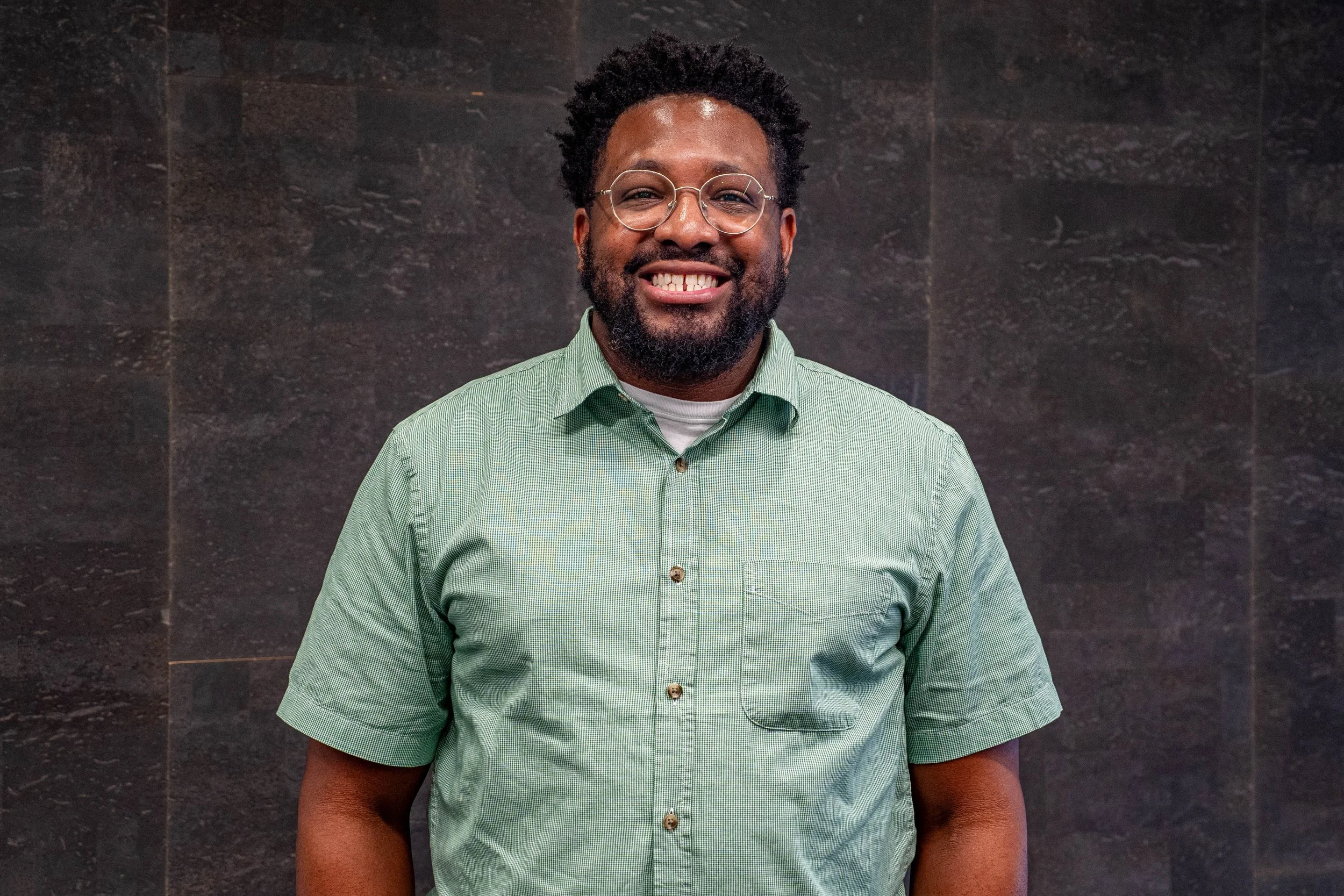 A woman with long dark hair smiling, wearing a black top and white jacket, sitting on a beige chair in front of a gray curtain.