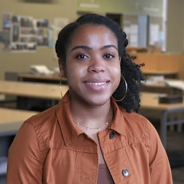 Portrait of a young African American woman smiling, wearing a blue blazer and white checkered shirt, against a gray background.