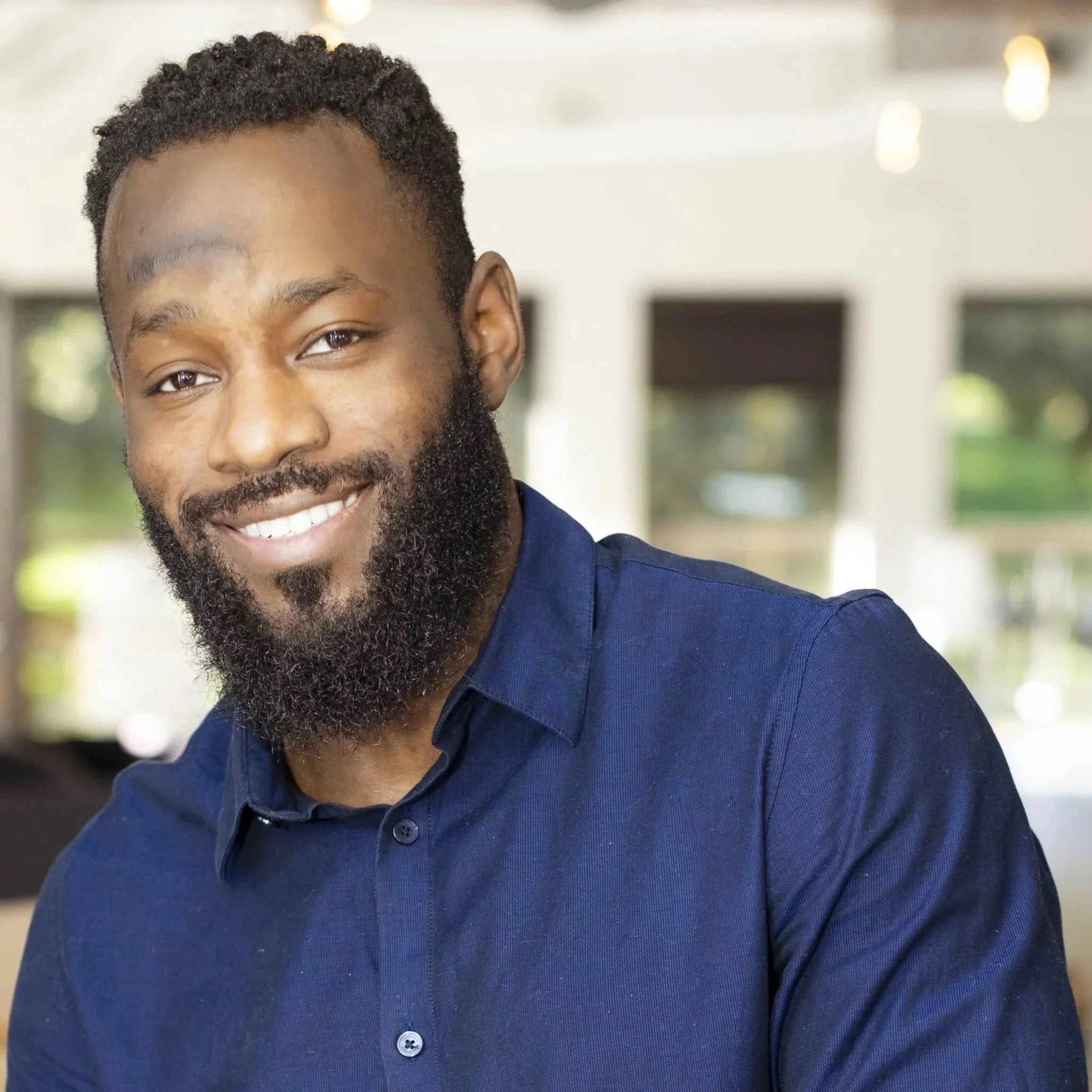 A professional man with glasses and a beard, wearing a navy blazer and pink shirt, smiling in an office setting.