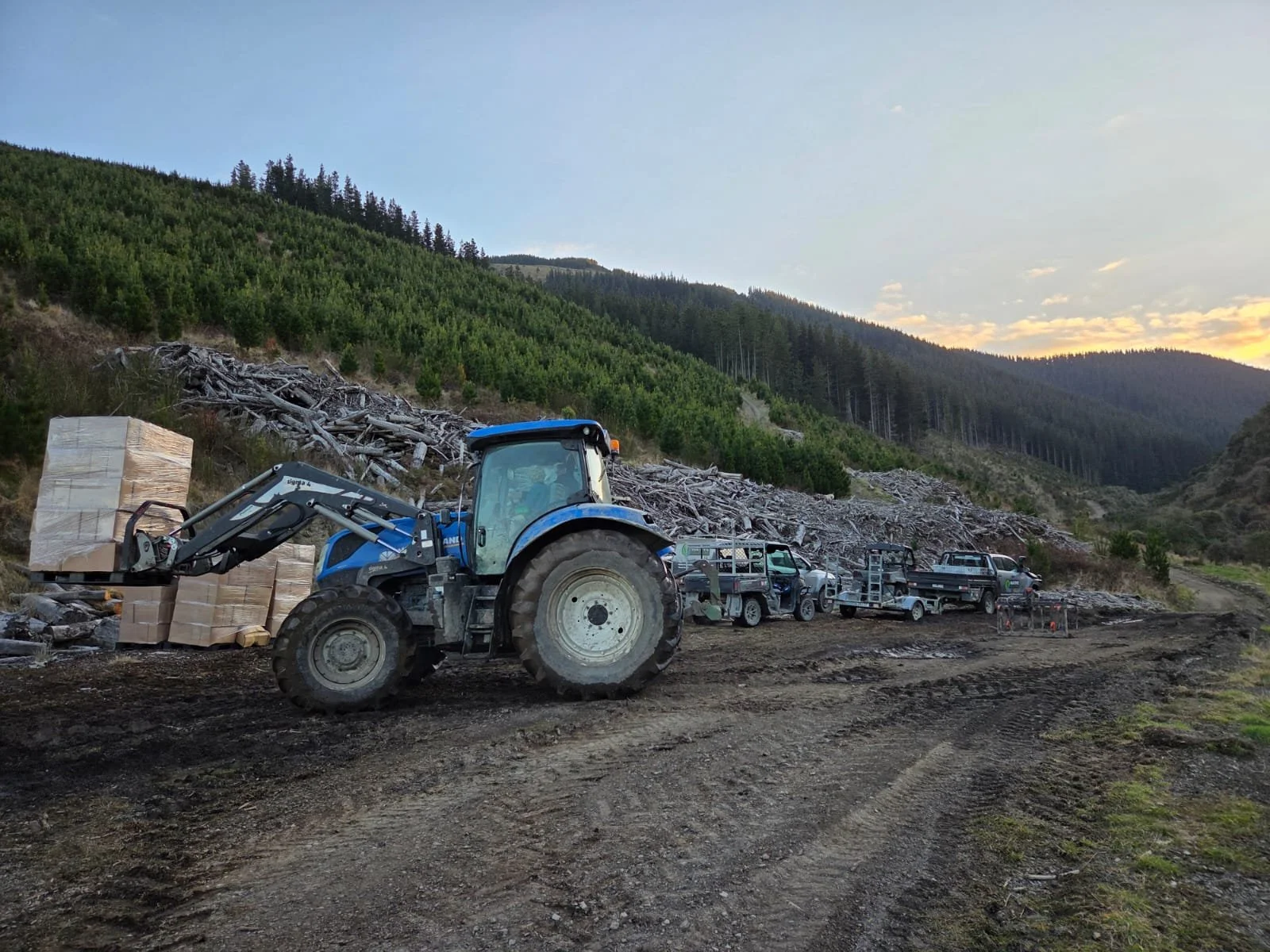 A construction site in a mountainous area shows a blue tractor with a front loader loading large cardboard boxes. Behind it, there are several pickup trucks and utility vehicles parked along the rough dirt road. The background features green forested hills and a partly cloudy sky at sunset.