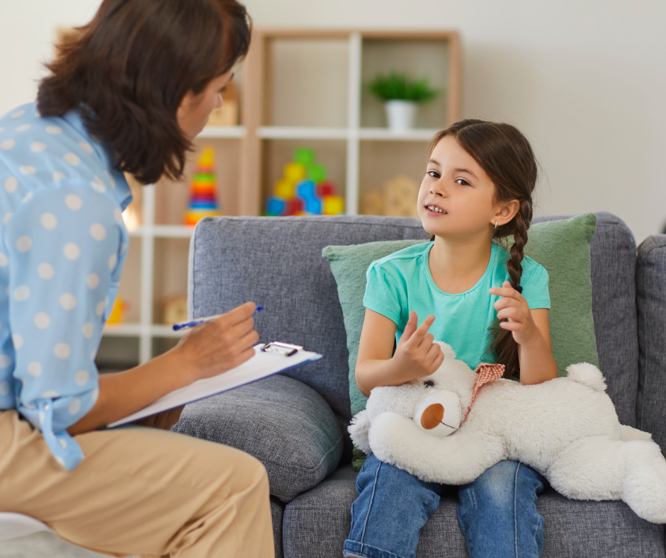 A young girl sitting on a gray couch holding a teddy bear, talking to a woman who appears to be a therapist or counselor, in a living room with a bookshelf in the background.