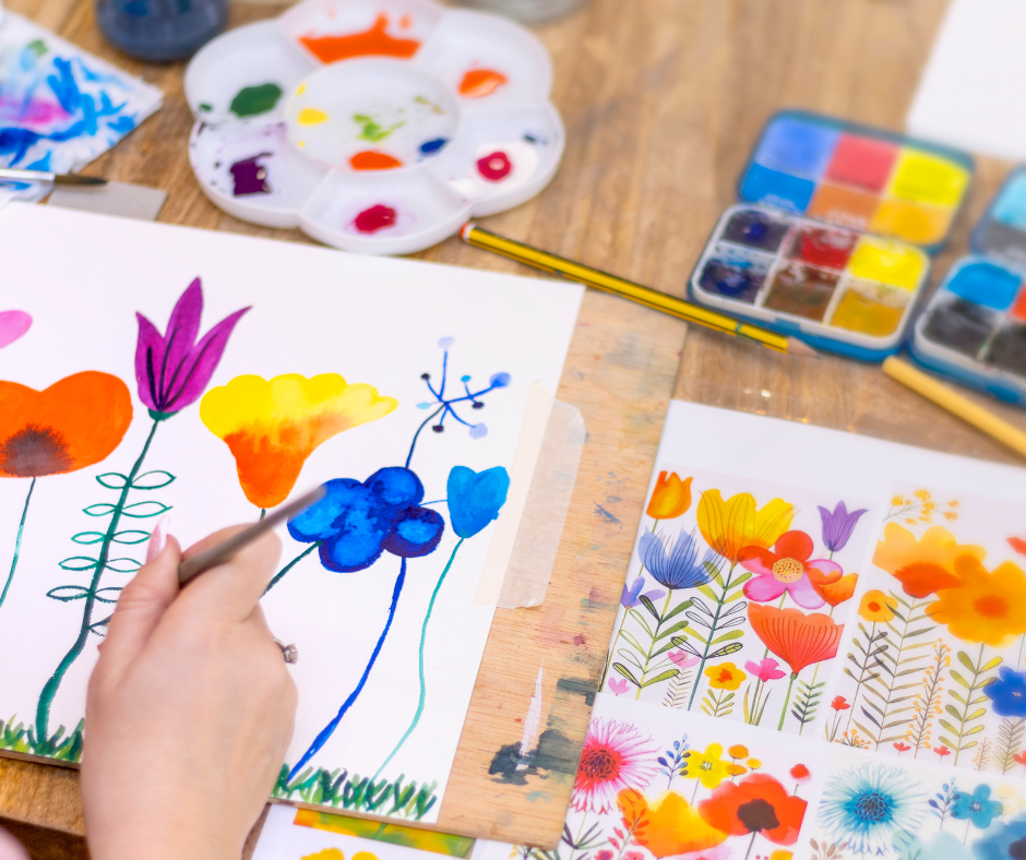 Person painting watercolor flowers on paper with various watercolor supplies and floral illustrations around.