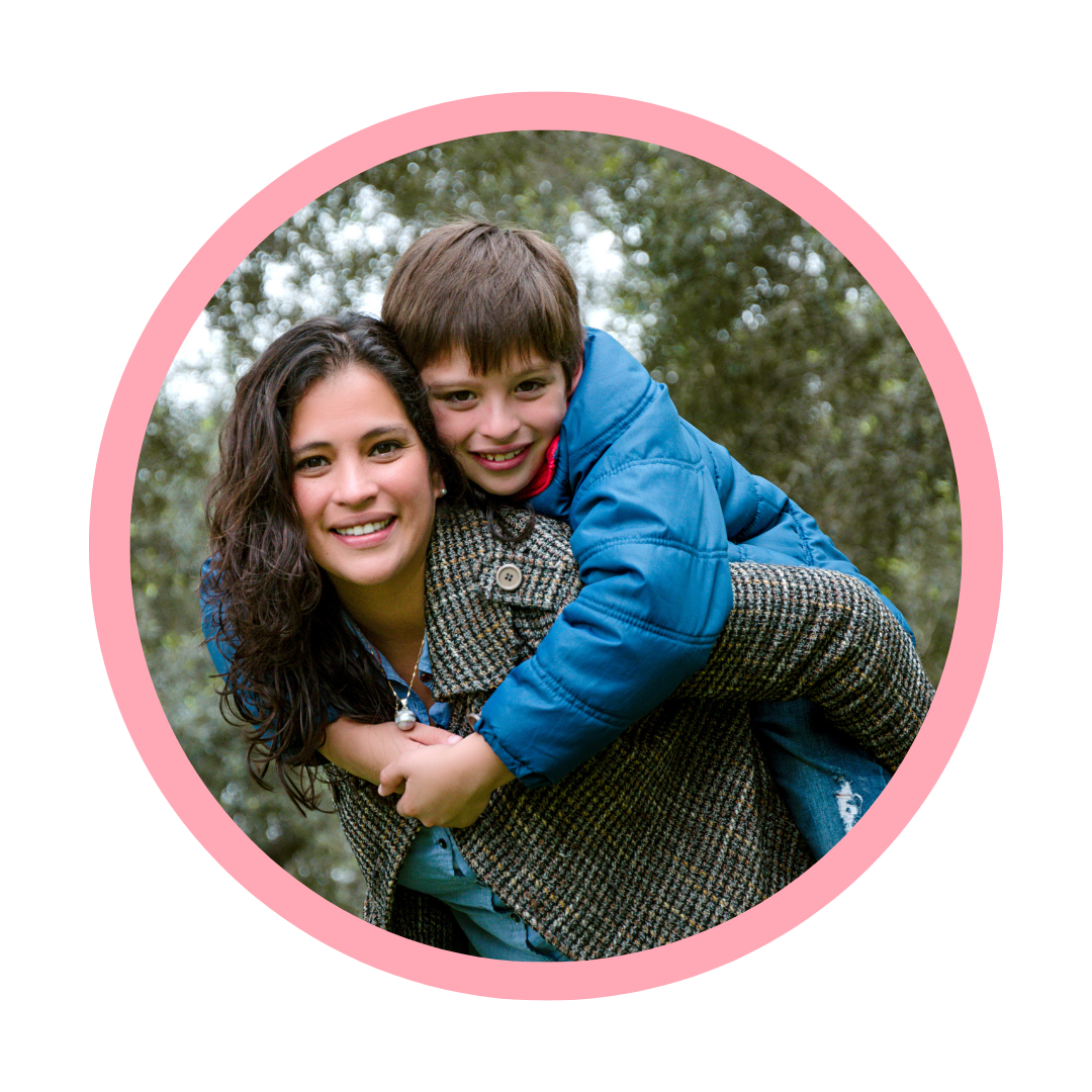 A woman with curly dark hair smiling while carrying a young boy on her back, both outdoors with trees in the background.
