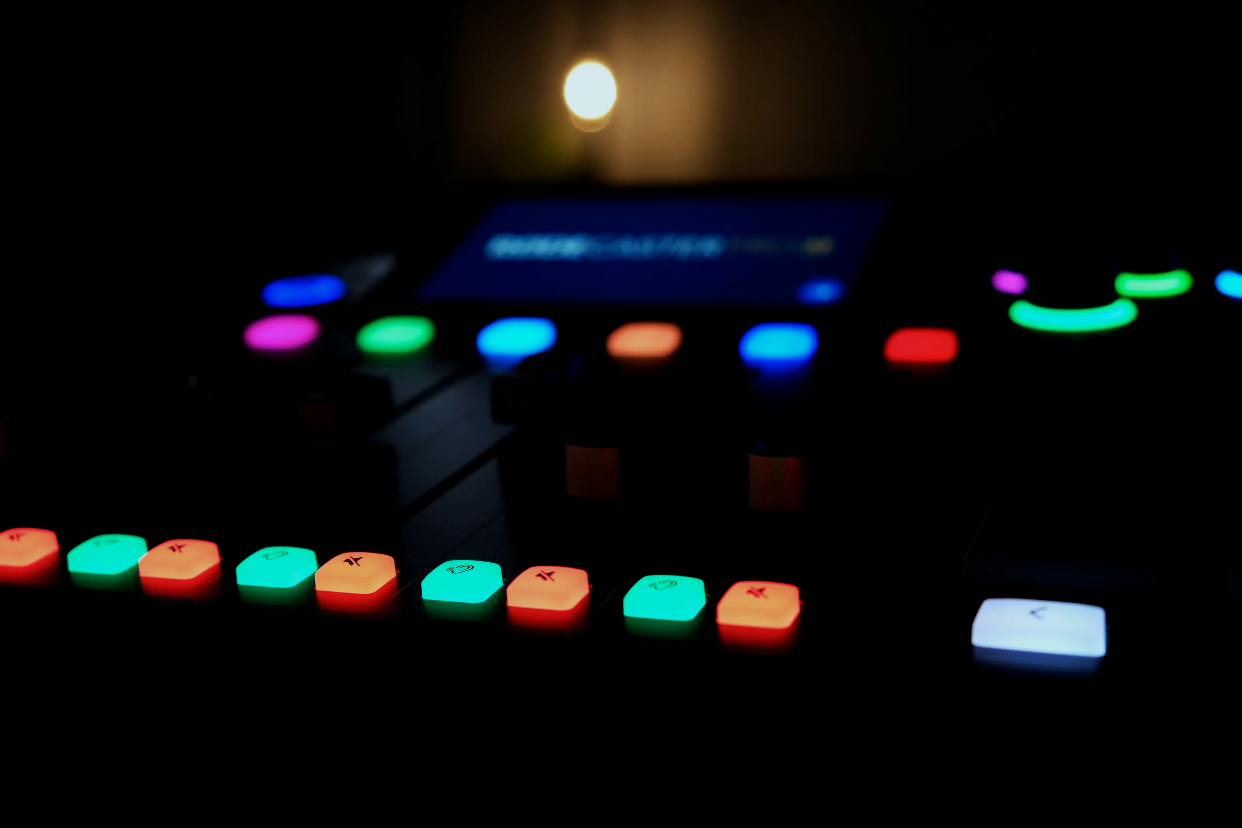 Dark photo of a music production device with multicolored illuminated buttons and touchscreen display in background.
