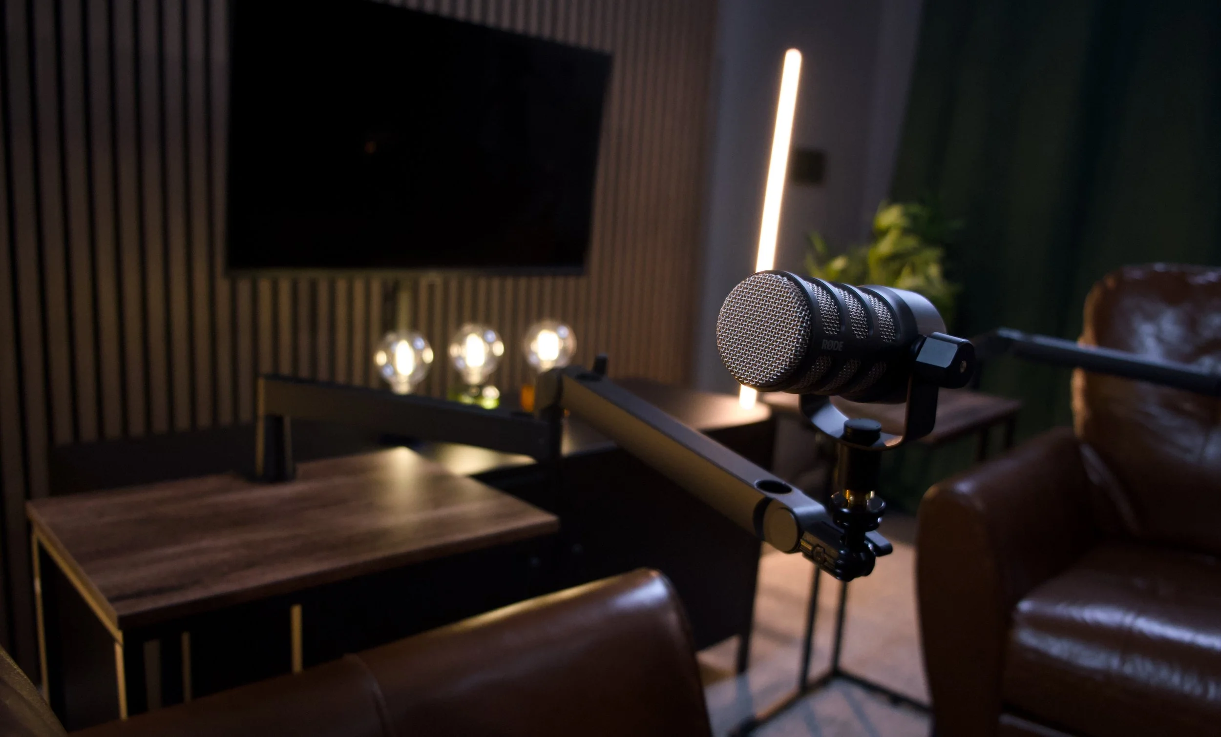 Close-up of a microphone mounted on a boom arm on a wooden desk, with a blurred background featuring a warm light source.