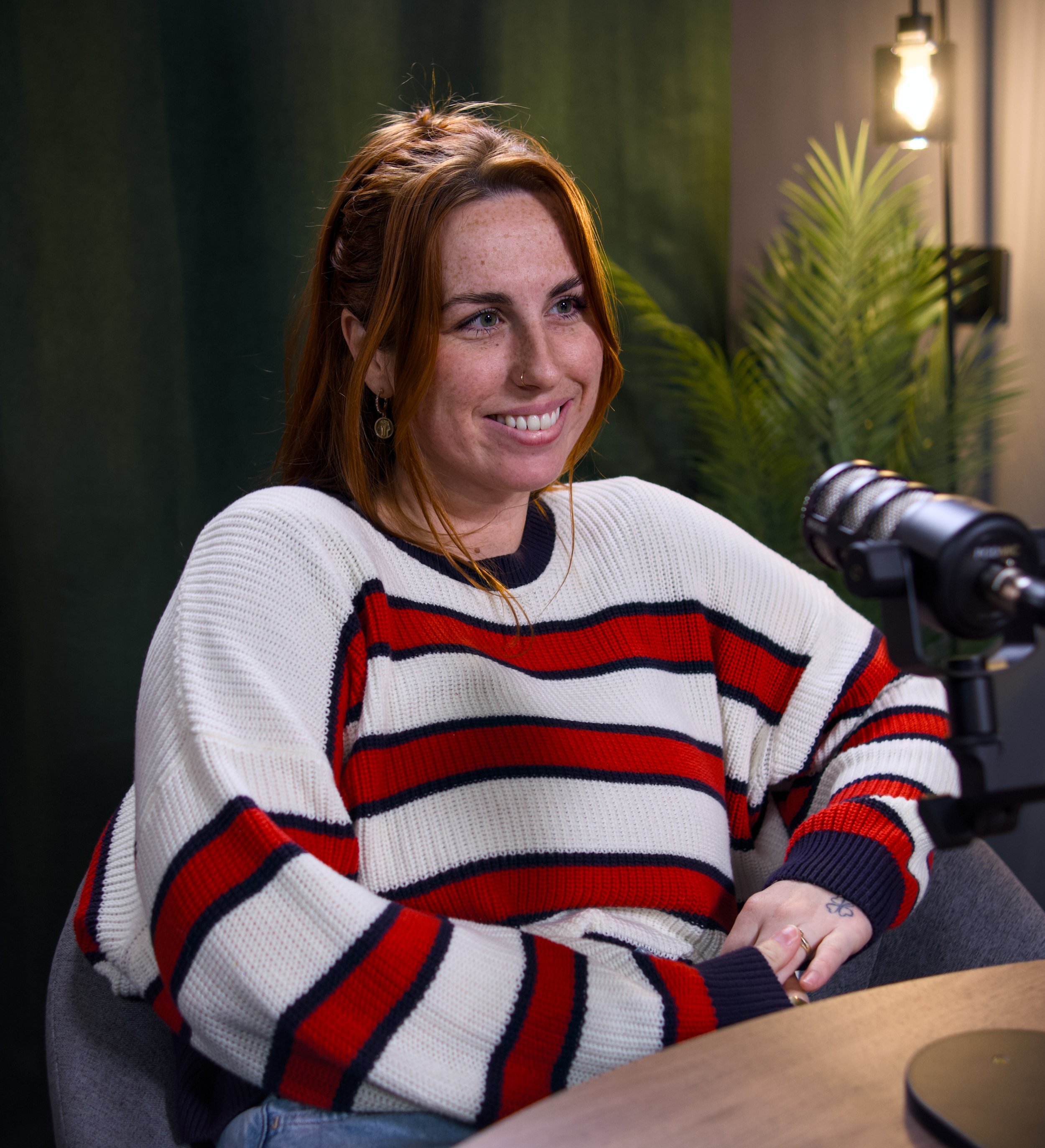A young woman with red hair, wearing a white sweater with red and navy stripes, is smiling and sitting at a table with a microphone in front of her. In the background, there is a green wall, a plant, and a warm light.