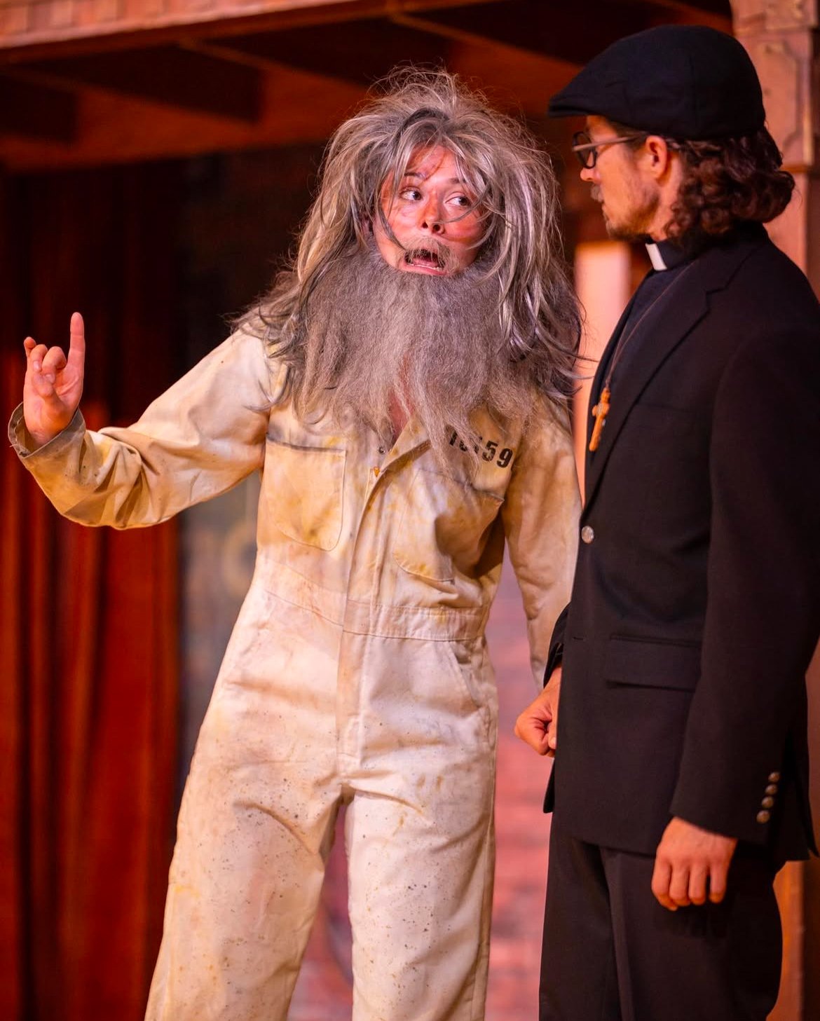 Emily Michelle Walton transformed as the character Barnardine in a prison uniform with a gray wig and false beard, speaking with Riley O'Toole as the Duke disguised as a friar in Measure for Measure at Montana Shakespeare in the Parks.