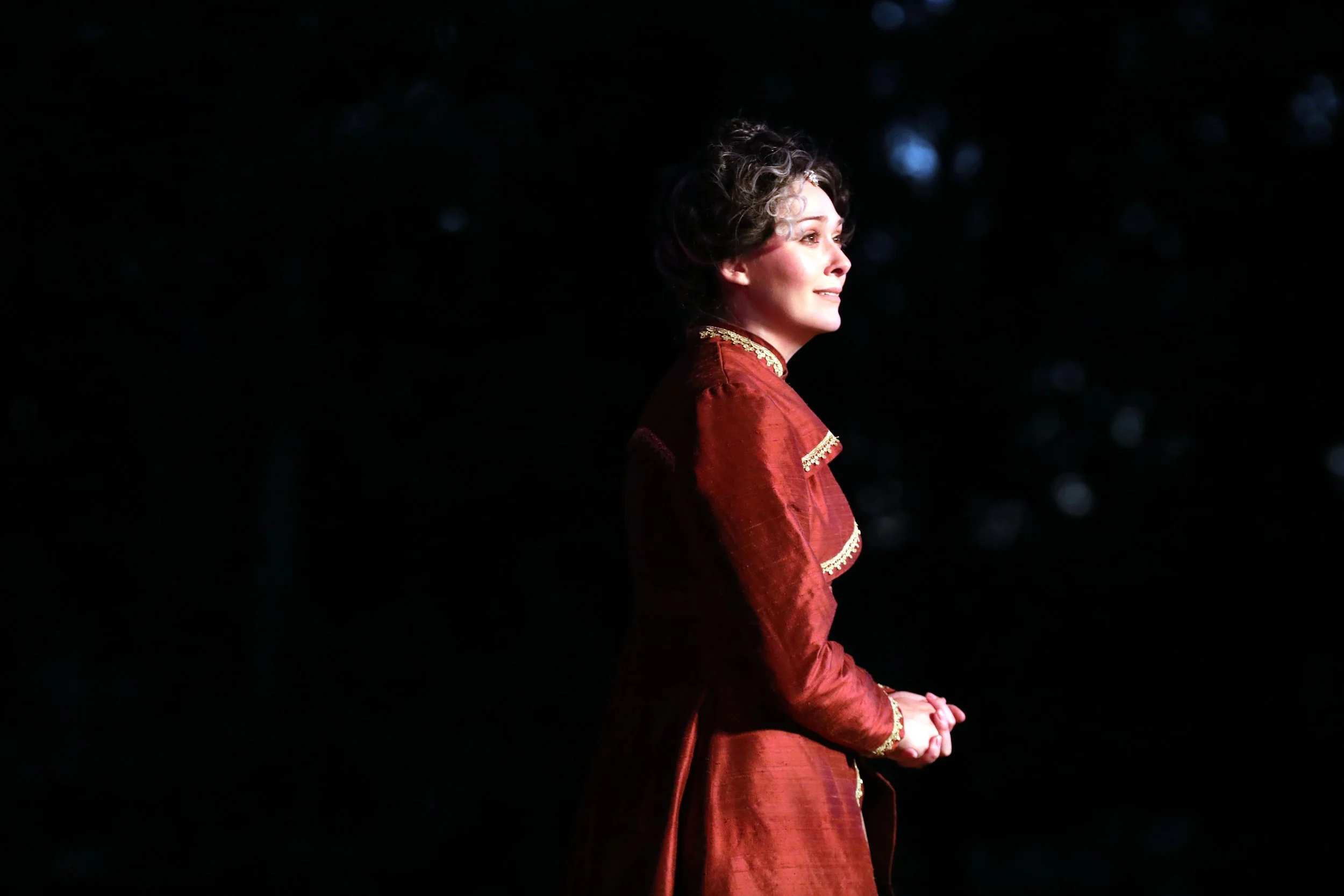 Emily Michelle Walton as Gertrude in Hamlet at Montana Shakespeare in the Parks, standing in profile in a red gown with gold trim against a dark background.